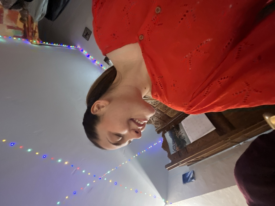 A young adult female, with brown hair styled in a ponytail, is captured in a close-up, low-angle shot within a room in Ancenis, France. She wears a red, long-sleeved top featuring eyelet patterns and three visible wooden buttons. Her head is angled downward and to the left, displaying a smile with teeth. The environment includes a light-colored ceiling and walls, illuminated by strings of multi-colored decorative lights (blue, green, red, yellow) extending across the ceiling and down a wall. In the mid-ground, a dark brown wooden screen or partition with intricate carved details is visible, upon which a calendar featuring a landscape image is affixed. A small, rectangular blue and white framed artwork hangs on a distant wall to the left. The lower-left foreground shows a partial view of a dark purple fabric object and a metallic gold-colored item.