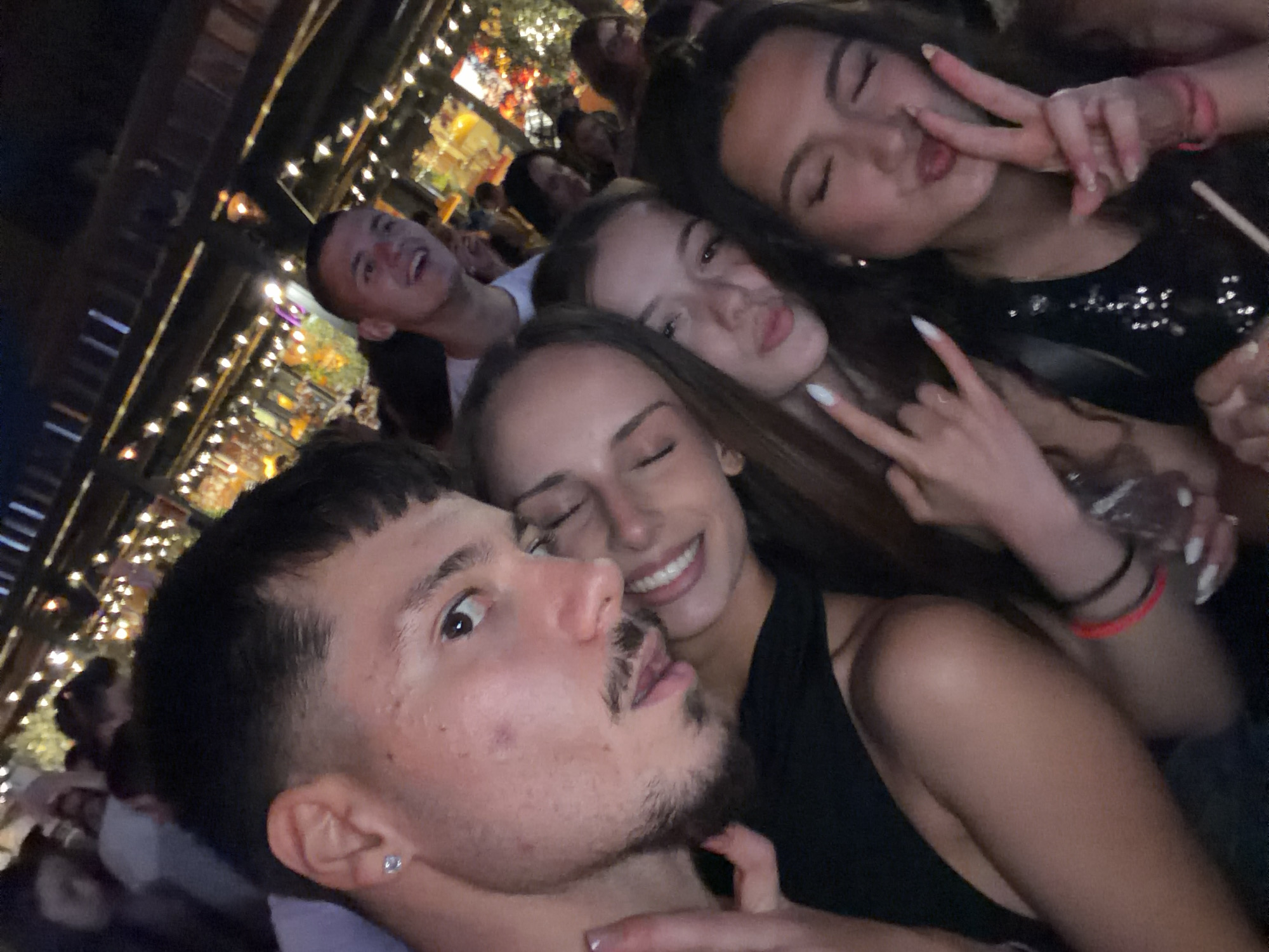 In this selfie, taken at a dimly lit outdoor venue, likely a bar or restaurant with string lights, five young adults are featured. The focus is on the faces of the individuals. The person closest to the camera, a young man with dark hair and a beard stubble, is looking directly at the lens. Beside him, a young woman with long brown hair is smiling and her face is tilted towards him. To their right, another young woman with long brown hair is making a kissing face. Behind them, a young man with short dark hair is visible, smiling and looking slightly upwards. Another young woman, partially obscured, is in the background, also looking upwards. The atmosphere appears festive and social. The location in the city of Sofia, Bulgaria, is not discernable from the image.