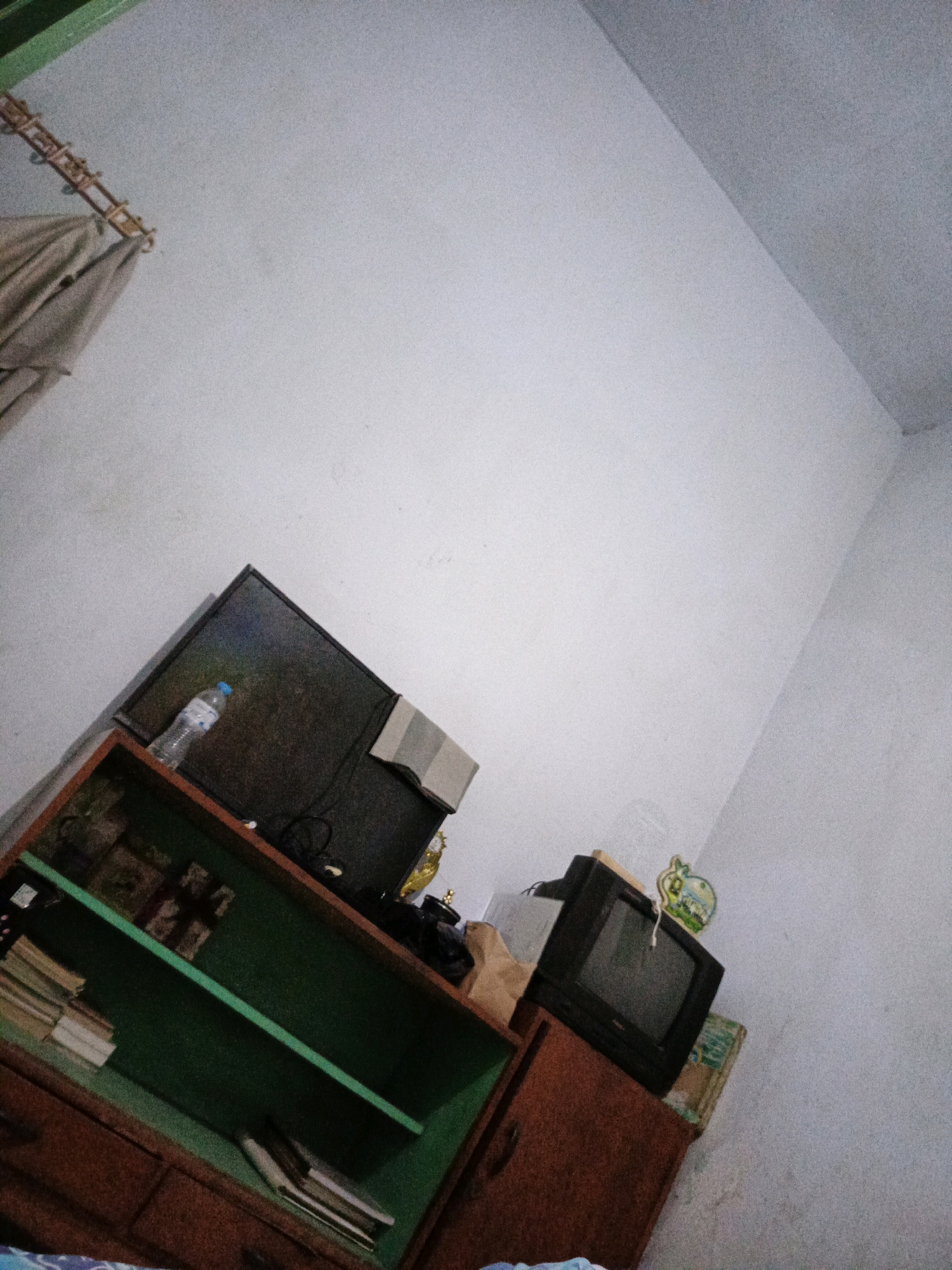 The image shows a dimly lit room with white walls. In the foreground, a wooden cabinet with green shelves is visible. The shelves contain stacks of books and some other items. On top of the cabinet, there are two televisions, one larger and black, and another smaller, older-model television. A plastic water bottle is placed on top of the larger television. To the left of the cabinet, a curtain hangs from a rod. The room appears to be a storage or living area. The scene is consistent with a residential interior in Sumbersari, Indonesia, likely a bedroom or living room given the presence of furniture and electronics.