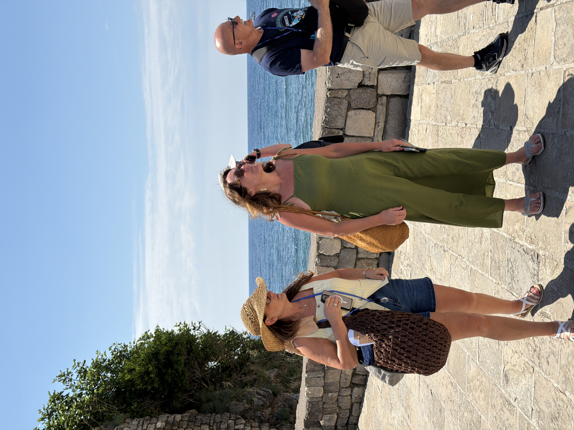 Three individuals are captured in an outdoor scene in Budva, Montenegro, likely sightseeing on a bright, sunny day. The image shows a paved stone pathway alongside a low stone wall, with the blue sea stretching into the distance under a clear sky with a few wispy clouds. Strong shadows on the ground indicate direct sunlight, suggesting midday or early afternoon.
From left to right, the subjects include:
A woman wearing a straw hat, a light-colored top, and denim shorts, holding a smartphone and carrying a brown woven bag. She is looking upwards and out towards the sea.
Next to her, another woman is dressed in a long, flowing olive green dress and sandals, also holding a smartphone and carrying a woven tan bag. She too is looking towards the horizon.
On the far right, a bald man wearing sunglasses, a dark blue t-shirt with "GREECE" prominently displayed on the front, khaki shorts, and sneakers stands with a black bag slung over his shoulder. He is also gazing out towards the sea.
All three appear relaxed, engaged with the scenic view, and dressed in casual summer attire suitable for warm weather and exploring a coastal location. Green foliage is visible behind the stone wall on the left, adding to the natural backdrop.