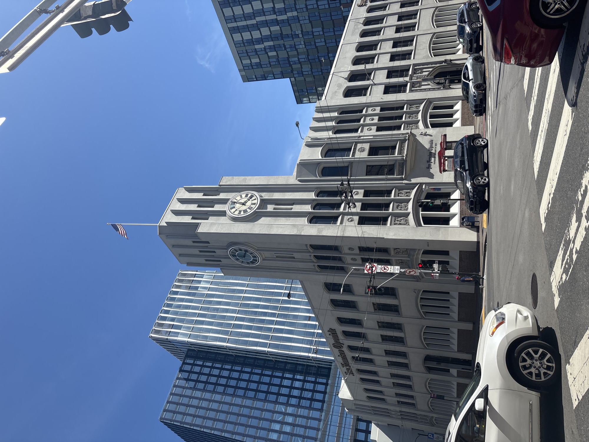 This is a low-angle, wide-shot photograph taken on a clear, sunny day in San Francisco, California, United States of America. The focal point of the image is the historic San Francisco Chronicle building, a striking Art Deco structure with a prominent clock tower. The building is made of light-colored concrete and features distinctive vertical fluting and arched windows. The American flag is flying from the top of the clock tower. To the left of the Chronicle building, two modern, tall glass skyscrapers are visible, reflecting the bright blue sky. On the right, another modern building with a dark, patterned facade is also visible.
In the foreground, a white SUV is parked or driving on the street, partially obscuring a crosswalk. Several other vehicles are also present on the street, including a silver sports car and a dark gray SUV. A red car is visible in the lower right corner of the frame. The street is paved with asphalt, and traffic signals and streetlights are present. A pedestrian can be seen in the distance, on the sidewalk near a traffic light. Traffic signs, including a "No Left Turn" sign and a sign indicating "MISSION," are also visible. The overall impression is a blend of historic and modern architecture characteristic of San Francisco.