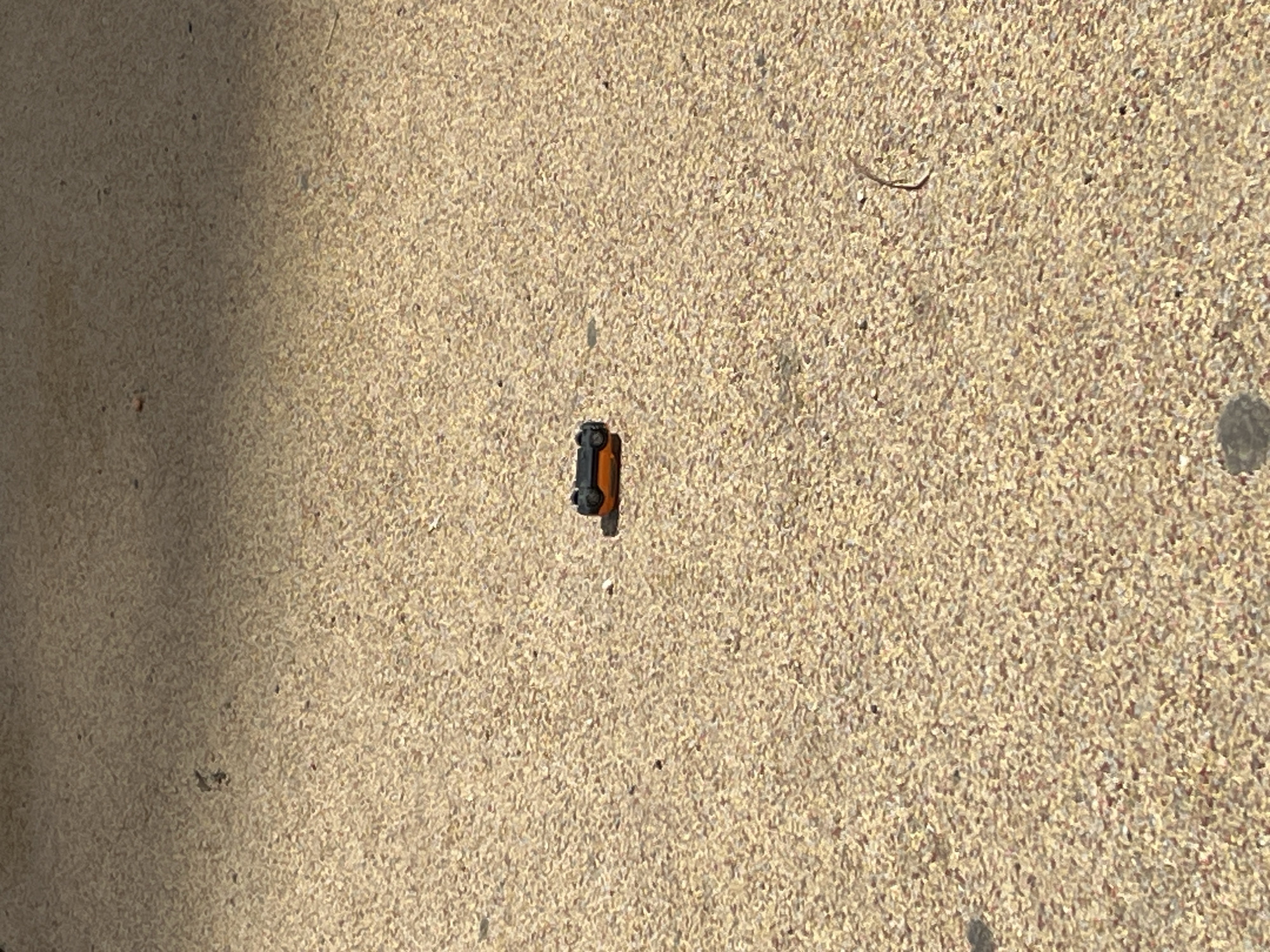 A small, orange and black toy car lies on a concrete surface. The background is a rough, light beige concrete. There is a dark shadow on the left side of the frame. The location and city cannot be determined.
