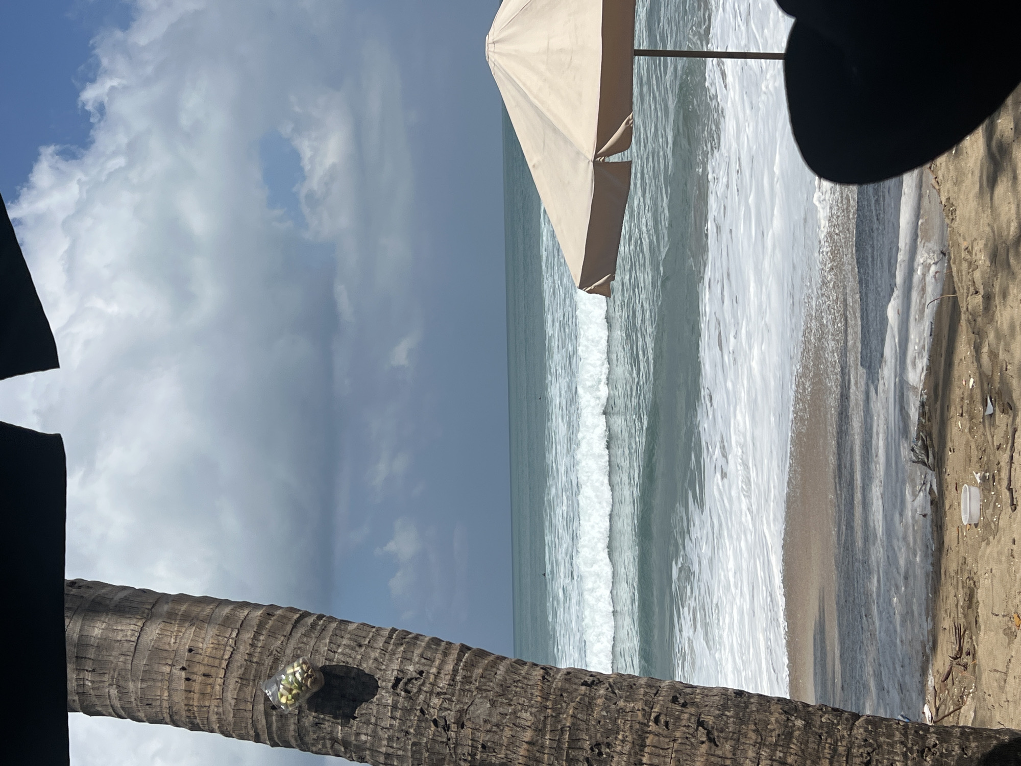 An outdoor shot captures a tropical beach scene in Kuta, Indonesia. In the foreground, a portion of a palm tree trunk is visible on the left, with a clear plastic bag containing small items, possibly snacks or offerings, tied to it. A beige beach umbrella stands on the right, angled towards the ocean. The ocean itself features turquoise water with white-capped waves rolling towards the shore. The beach sand is light brown, with scattered debris visible, including a white plastic container. The sky above is a mix of blue and heavy grey clouds. The overall impression is a slightly overcast day at a popular beach destination.