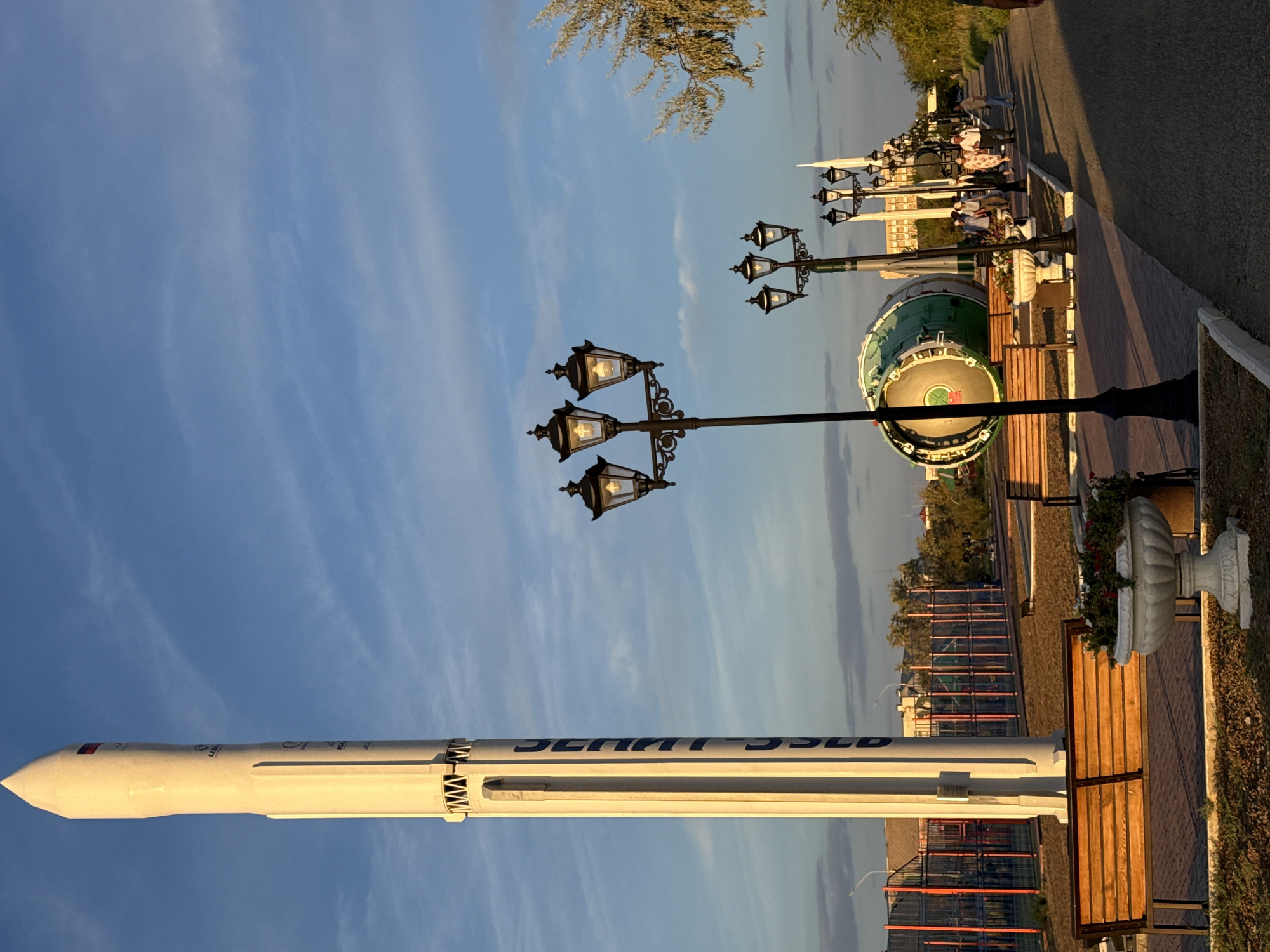 The image depicts a park area in the city of Baikonur, Kazakhstan, likely a monument or memorial related to space exploration. Dominating the left side of the frame is a tall, white rocket mockup, adorned with blue lettering and a small flag emblem near its apex. In the middle ground, a large, green, cylindrical object, possibly a rocket stage or a booster, rests horizontally on a stand. A paved walkway extends from the foreground into the distance, lined with ornate, multi-headed street lamps. Several people are walking along the pathway, dressed in casual attire. Benches are placed along the walkway, some with planters containing flowers. In the background, buildings, fences, and trees are visible, suggesting an urban park setting. The sky is a clear blue with wispy clouds, and the golden hour sunlight casts long shadows across the scene, indicating it is late afternoon or early evening.