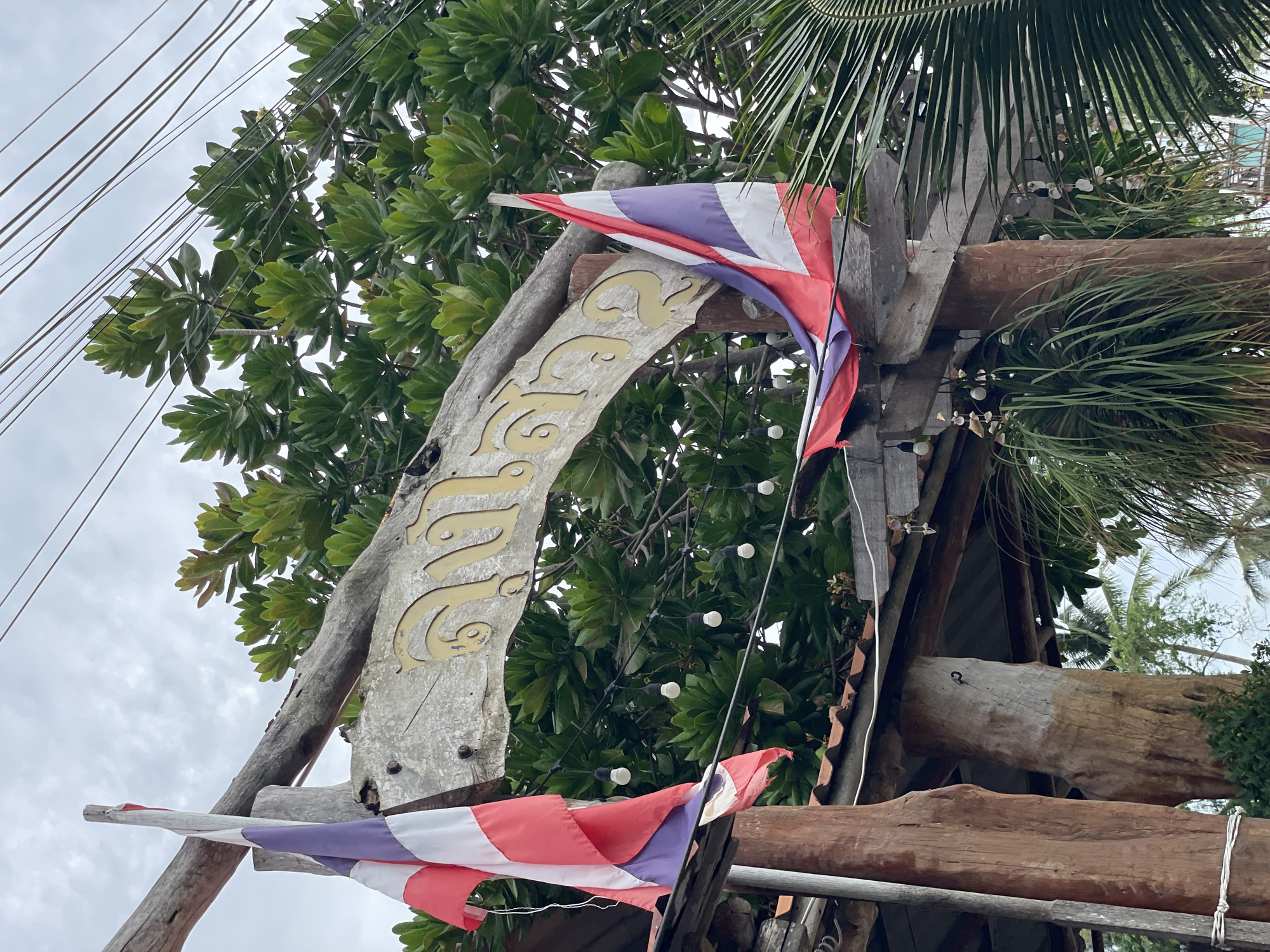 This is a medium shot of a wooden archway with Thai flags and a sign in Thai script. The archway is constructed from weathered, rough-hewn logs and planks. Two Thai flags, one on the left and one on the right, are draped over the structure. The left flag is attached to a vertical wooden pole and extends to the left. The right flag is attached to the top horizontal beam and hangs down over the arch. The sign, which appears to be made of weathered wood with gold-colored raised lettering, is mounted on the horizontal beam of the archway. The Thai script on the sign is legible but the exact words are not immediately discernible without translation. Behind the archway, a dense canopy of green leaves from a large tree is visible, with some electrical wires running above it. String lights with visible bulbs are also hanging from the underside of the archway. The overall scene suggests an entrance to a establishment or a significant point of interest in a tropical or coastal environment, likely in Ban Na Chom Thian, Thailand, given the presence of Thai flags and vegetation. The sky is overcast.