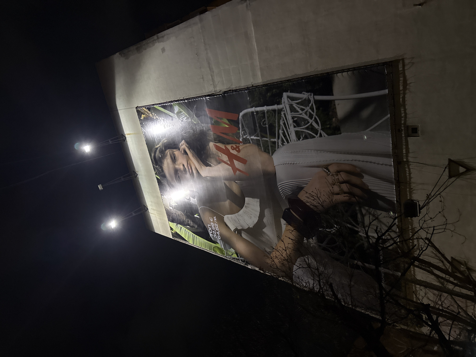 The image shows a large H&M billboard mounted on the side of a building, captured at night in Varna, Bulgaria. The billboard features a model in a white outfit against a blurred background with green foliage and a white wrought-iron chair. The H&M logo is visible in red. Streetlights illuminate the billboard. A bare tree partially obscures the lower right corner of the billboard. The building's facade is plain and appears to be concrete or stucco. The sky is dark.
