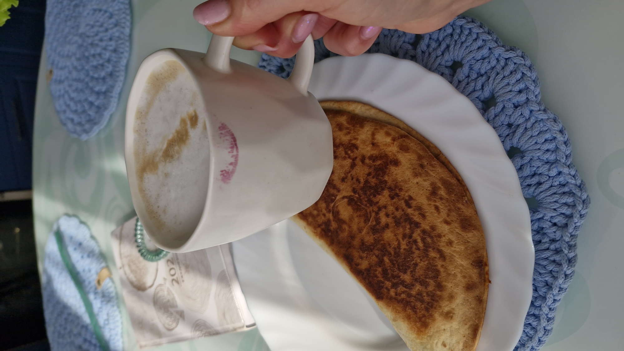 A person's hand holds a white ceramic mug with a visible lipstick mark, filled with a light brown beverage, presumably coffee. A white plate below holds a folded, golden-brown flatbread or pancake. The plate is on a light blue crocheted placemat. A portion of a calendar is visible in the lower left, as is a small blue crocheted circle and green ring. The scene appears to be set indoors on a table. The exact location in Otchor, Russia, is undeterminable from the image.