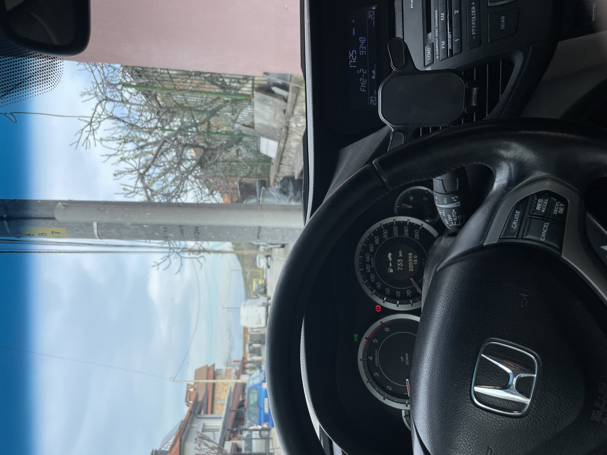 The image shows the interior of a car, specifically the dashboard and steering wheel of a Honda vehicle. The car is stationary, as indicated by the speedometer reading "0". The odometer shows "220398" km and the trip meter displays "733 km". The radio is tuned to FM2-2 with a frequency of 93.40, and the time is 17:25. The outside temperature is 20 degrees Celsius. A mobile phone holder is attached to the dashboard.
Outside the car window, there is a view of a residential area on what appears to be a street, with buildings and trees visible. The presence of a street sign suggests a city location.