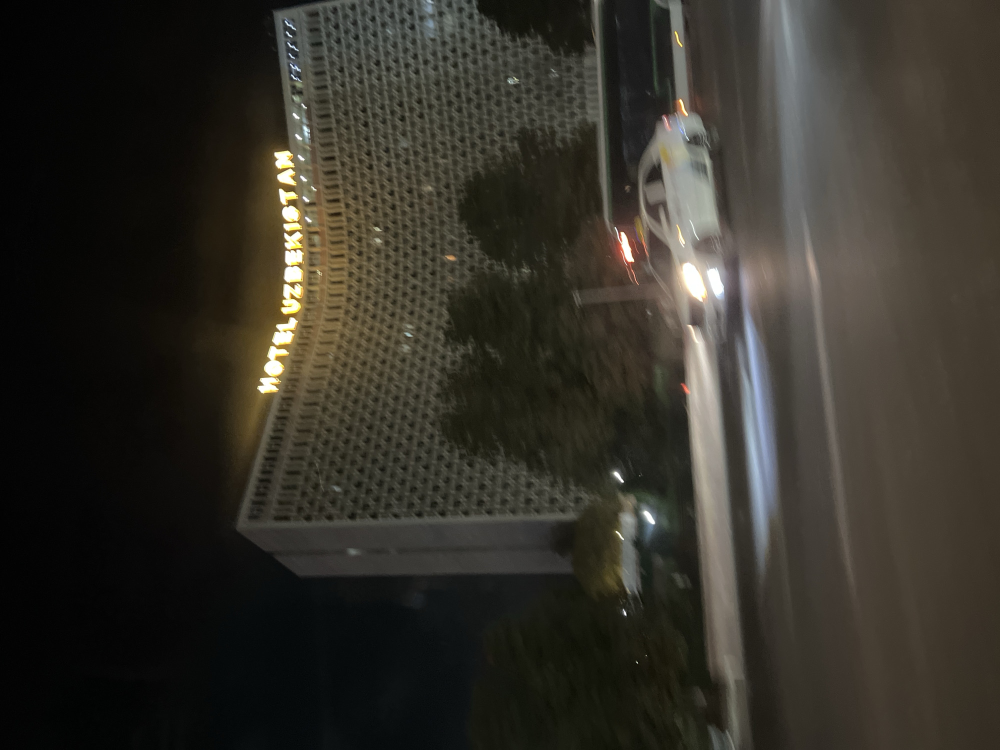 The image depicts a nighttime scene on a city street in Kashgar Mahalla, Uzbekistan. A large, modern building with "HOTEL UZBEKISTAN" illuminated in yellow neon lights on its facade dominates the background. The hotel has a distinctive curved design with a repeating geometric pattern on its exterior. In the foreground, a blurred white car, possibly a taxi, is driving on the wet street, its headlights illuminating the road. A darker, longer vehicle, likely a bus, is visible behind the car. Trees line the street between the hotel and the road. The overall impression is one of motion and urban nighttime ambiance, with the car and bus showing motion blur, indicating they are in transit.