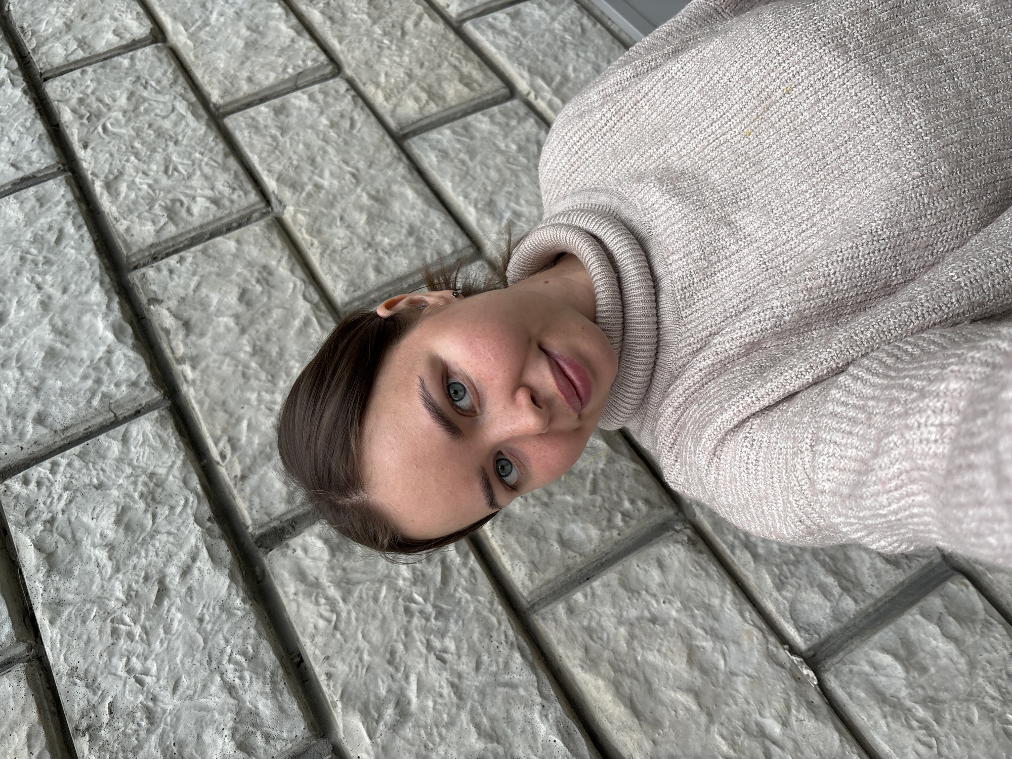 A young woman with blue eyes and dark hair is lying on a brick or stone paved surface. She is wearing a light-colored, possibly beige or light grey, turtleneck sweater. The pavers are grey and textured, arranged in a rectangular pattern. The picture appears to be taken outdoors. The location is Русский Вожой, Russia.