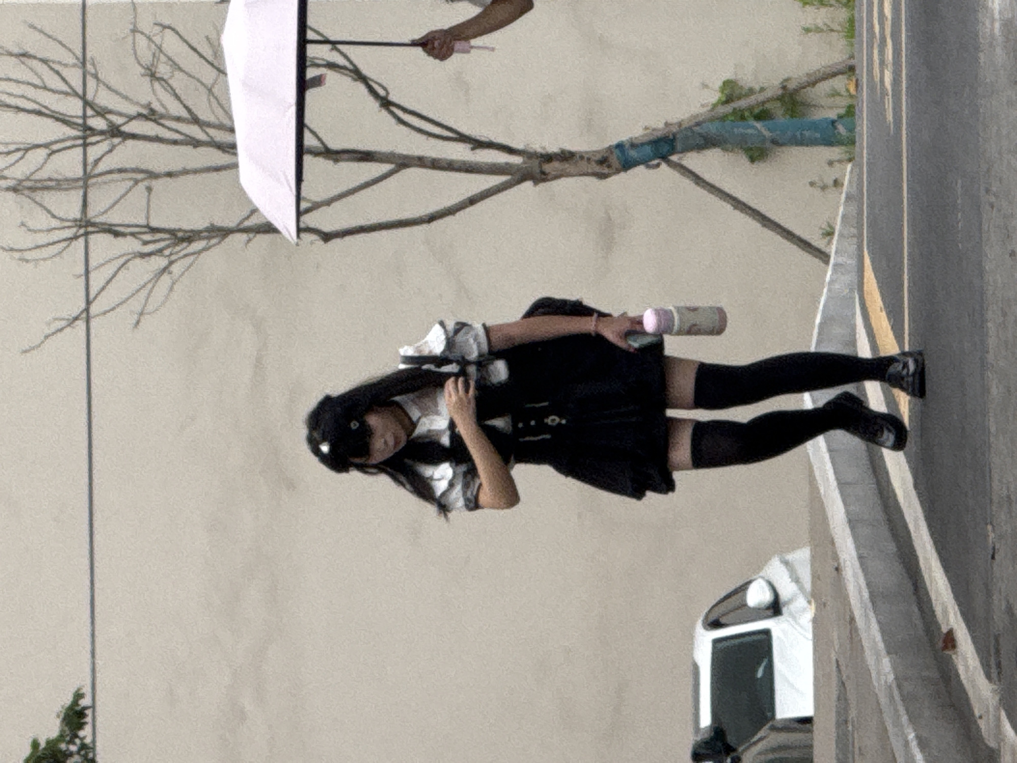 A young woman in a dark skirt, white collared shirt with a dark tie, and black stockings stands on a city street beside a concrete curb. A white commercial van is parked nearby. The woman appears to be holding a pink-labeled beverage container. An umbrella is held by a hand at the upper left of the frame. A bare tree branch extends into the scene from the left. The location is in Jiangjin, China.