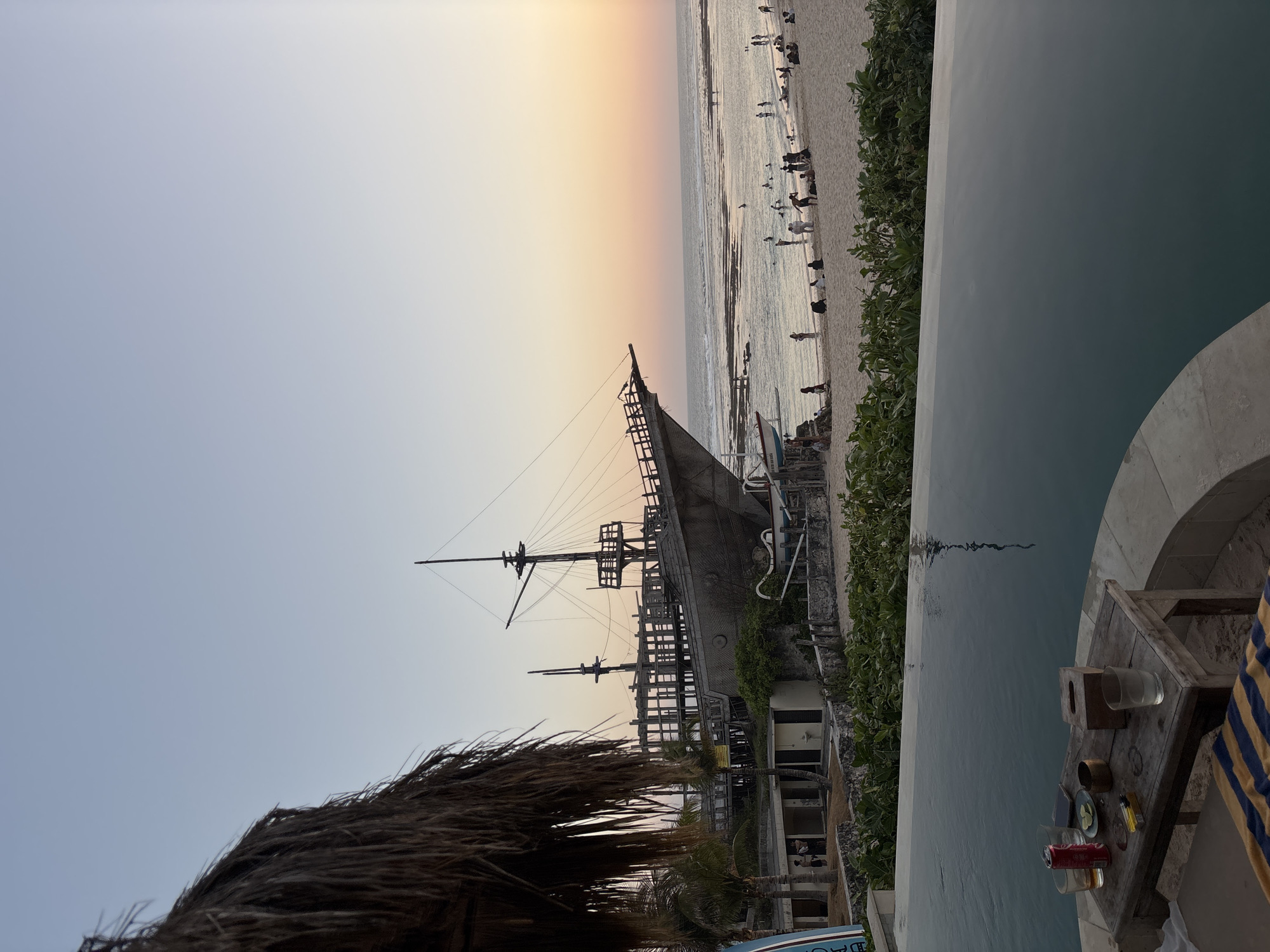 This image captures a serene beach scene in Kuta Selatan, Indonesia, during the golden hour. In the foreground, a portion of an infinity pool reflects the soft hues of the twilight sky. Beside the pool, a rustic wooden table holds a few drinks, a pack of cigarettes, a lighter, and a tissue box, suggesting a moment of relaxation.
Beyond the pool, a sandy beach stretches towards the ocean, dotted with numerous people enjoying the late afternoon. Some are wading in the shallow water, while others are seated or standing on the sand, likely observing the sunset.
Dominating the mid-ground is a large, weathered, ship-like structure, possibly a restaurant or attraction, positioned near the shoreline. The structure has the appearance of a historical sailing vessel with multiple decks and masts. Adjacent to it are some buildings, likely part of the same establishment.
The ocean stretches to the horizon, with gentle waves rolling onto the shore. The sky transitions from a pale blue at the top to soft shades of orange and pink near the horizon, indicating the sun is setting. Lush green vegetation lines the area between the beach and the structures. The overall atmosphere is tranquil and picturesque, highlighting a popular tourist destination in Kuta Selatan.