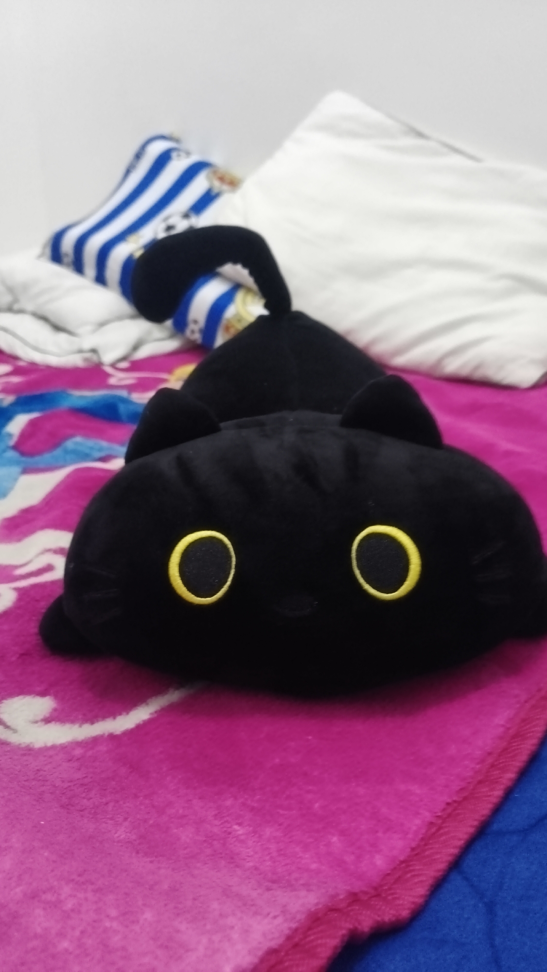 A black stuffed cat with large yellow eyes and black ears lies on a pink blanket. Behind the cat, a white pillow and a blue and white striped pillow are visible. The background is a white wall. The image appears to be taken in a bedroom. There is no indication of the location being Betung, Indonesia.