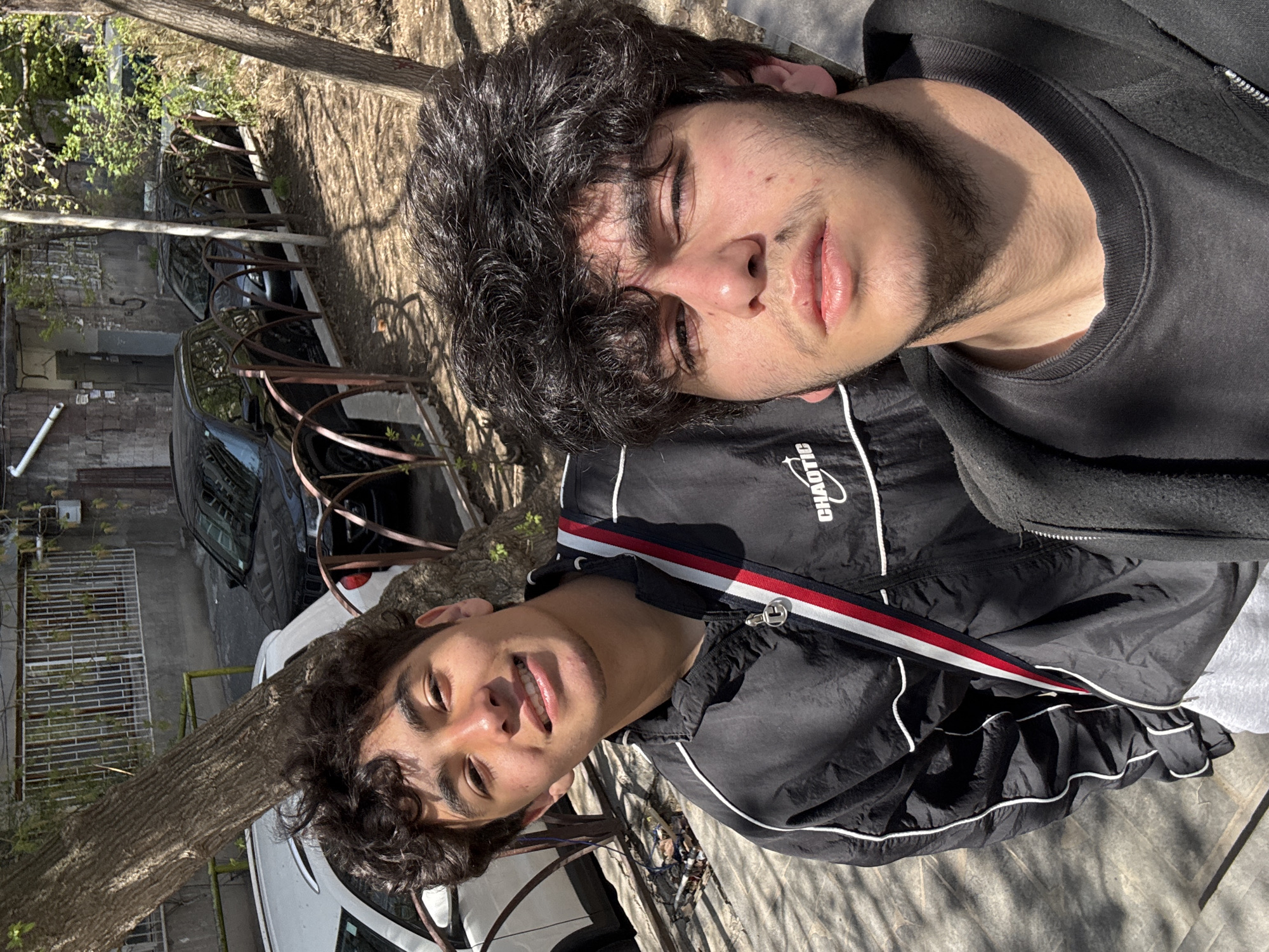 The image shows two young men in Yerevan, Armenia, seemingly posing for a selfie outdoors. One man is wearing a black "CHAOTIC" jacket with white and red stripes, while the other wears a black sweatshirt. They are standing next to a tree. In the background are parked cars and a building with brick walls and a metal gate. The setting appears to be a street or courtyard within the city.