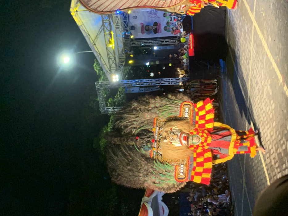 The image shows a nighttime performance in the city of Kecamatan Batu, Indonesia, specifically at the coordinates -7.867 latitude and 112.514 longitude. The image is of a street performance featuring a costumed dancer with a large, intricately-designed headdress and mask. The headdress is adorned with feathers, resembling a peacock, and there is a sign on the mask that reads "PONO ROGO". The dancer is wearing a red and yellow checkered garment with black accents and is dancing barefoot on a paved street, illuminated by a single streetlight overhead. The surrounding environment is dimly lit, and a crowd of spectators can be seen in the background, gathered in front of a large stage. The stage is covered with a white tent-like structure and is equipped with lighting and sound equipment. There is a large banner behind the stage with an image of a man and a smaller, smaller banner featuring a group of people. The dancer is performing in front of the stage. The background is filled with trees and a large, white tent-like structure.
