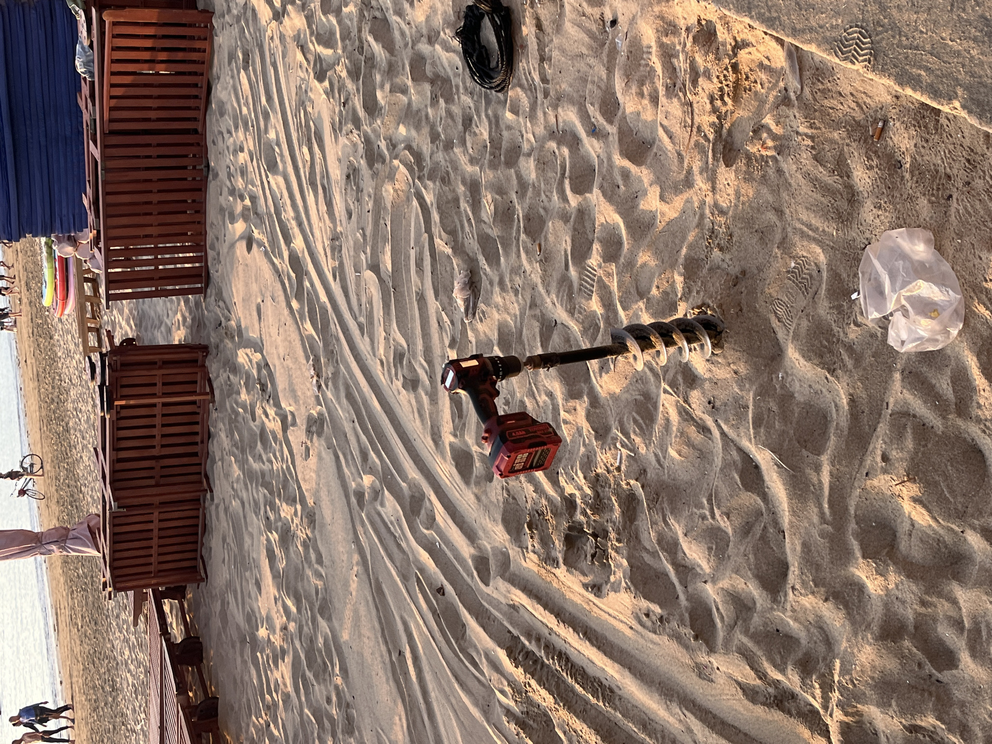 This image, likely taken in Bac Nha Trang Ward, Vietnam, depicts a sandy beach. A power drill with an attached auger bit sits on the sand. A plastic bag lies near the drill. A black cord is also visible on the sand. Along the left edge of the image are wooden structures, possibly beach chairs or sunbeds. Figures of people are visible in the distance near the water.
