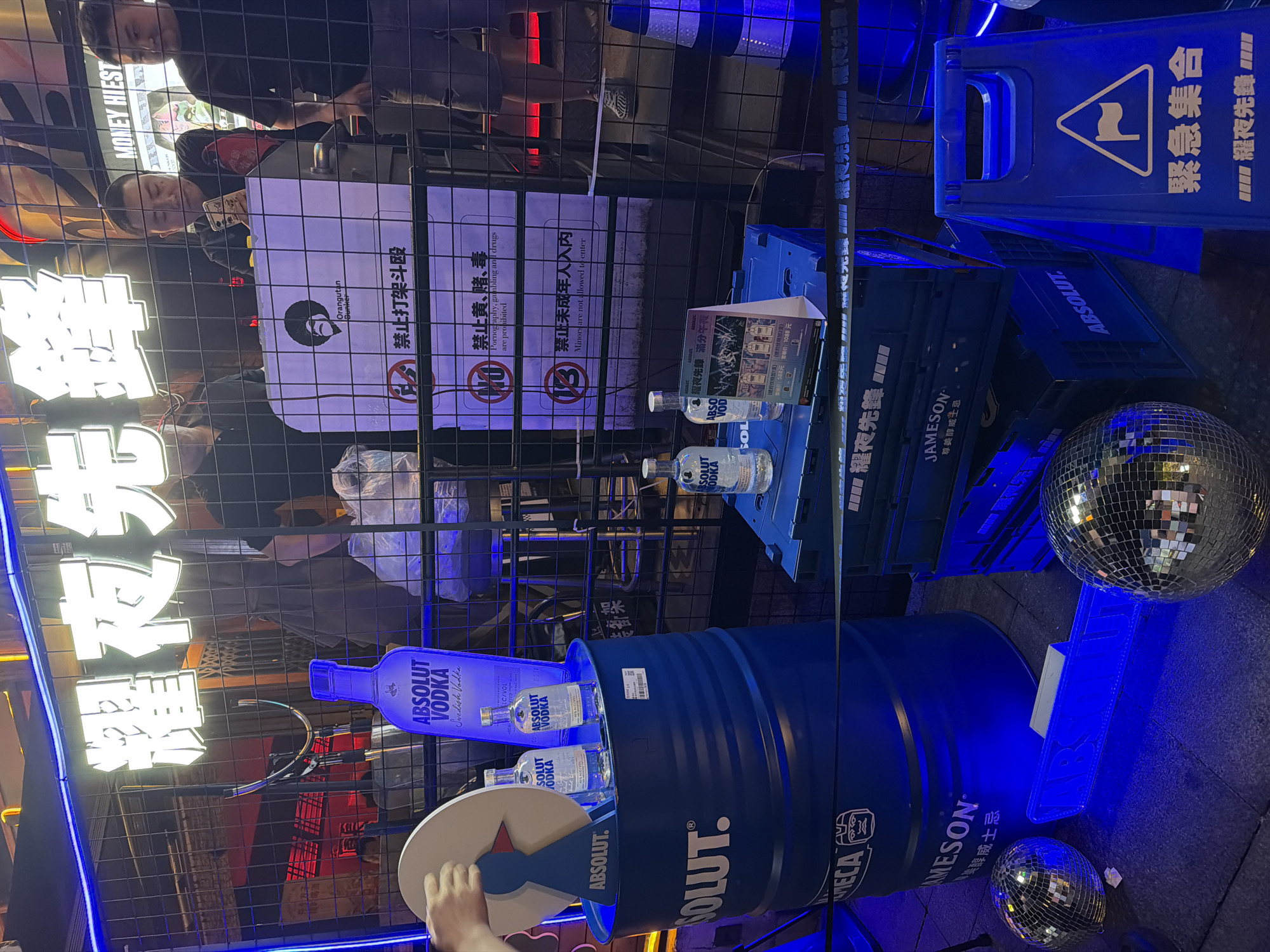 The scene depicts a nighttime outdoor display, likely in the Liangjiang area of China, judging from the Chinese writing present. The display is dominated by branding for ABSOLUT Vodka and Jameson Whiskey. An illuminated sign in Chinese characters is visible at the top left. A large blue metal barrel, marked with "ABSOLUT" and "JAMESON," serves as a base for multiple ABSOLUT Vodka bottles. A light-up blue ABSOLUT Vodka bottle replica lies horizontally on the barrel. Nearby are blue plastic crates branded with "ABSOLUT" and "JAMESON." A disco ball is placed on the ground beside the crates. A blue "Wet Floor" sign is also present. Behind the display, a black metal mesh fence is visible with signs containing Chinese text and prohibition symbols for fighting, gambling, and underage drinking. People are standing behind the fence, possibly observing the display. The overall lighting is predominantly blue, with neon lights highlighting the branding. A hand is visible near the barrel, possibly interacting with the display.