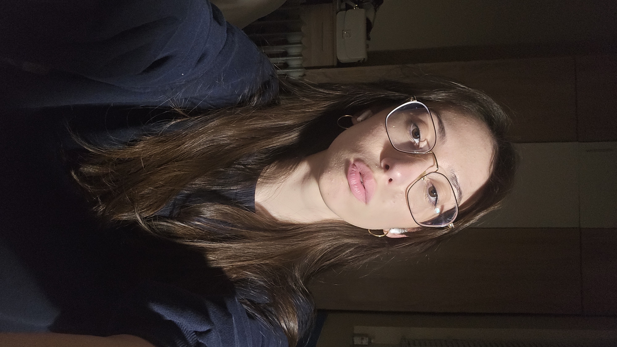 A woman is captured in a selfie taken indoors, possibly in a private residence or apartment in Stara Zagora, Bulgaria, given the absence of external landmarks. The woman has long, brown hair, and wears glasses with gold frames. She is dressed in a dark-colored shirt. Her face is prominent, revealing fair skin and light pink lips. The lighting is subdued, with a noticeable shadow partially obscuring the right side of her face. The background is blurred and out of focus, but elements resembling a cabinet, a white purse, and what appears to be a radiator can be discerned. She is wearing golden hoop earrings.