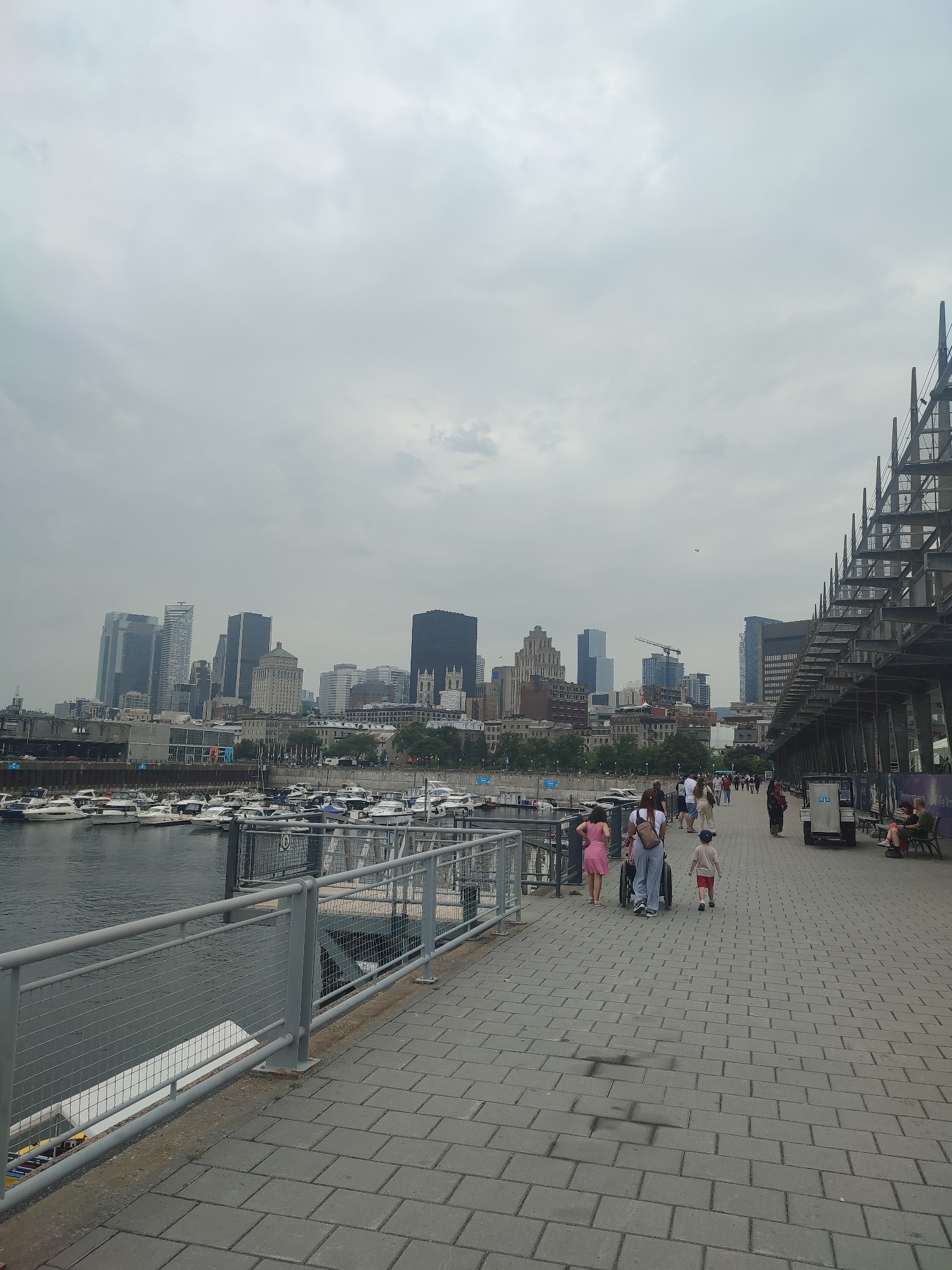 The image captures a waterfront promenade in Montreal, Canada, with the city's skyline visible in the background. The promenade is paved with gray cobblestones and lined with a metal railing separating it from a marina filled with numerous docked boats. On the right side of the promenade, a modern, angular structure with metal supports dominates the scene, suggesting ongoing construction or a contemporary architectural feature. Several people are walking along the promenade. In the foreground, a woman pushes a stroller with a child walking beside her. Further down the path, other individuals and groups are seen strolling. The sky is overcast, casting a diffused light over the scene. The overall impression is that of a busy urban waterfront on a cloudy day.