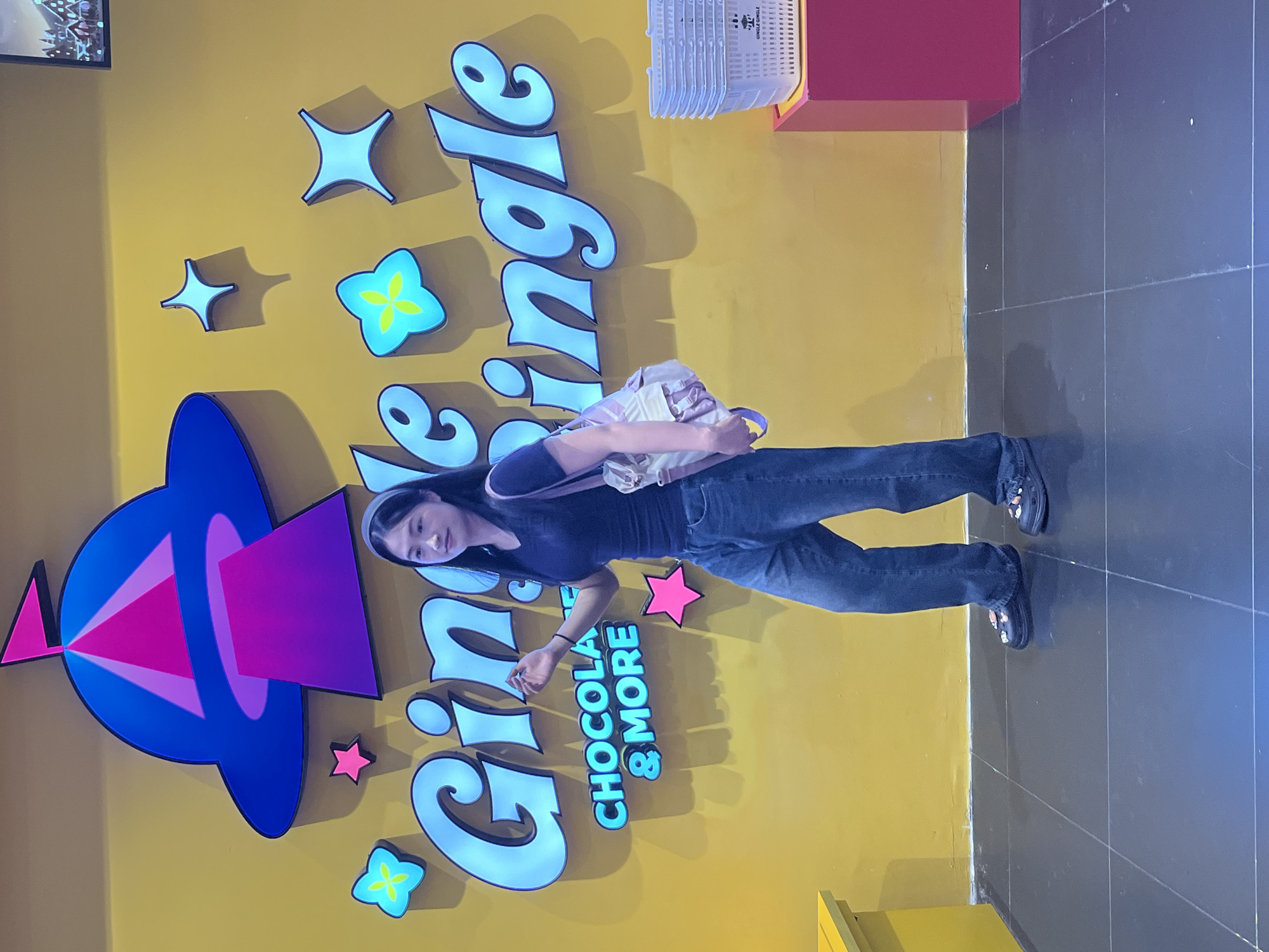 A young Asian woman stands in front of a yellow wall with a large, neon sign that reads "Gingle Gingle" in blue letters. Above the sign is a depiction of a UFO with a pink beam of light. The woman is wearing a dark t-shirt, baggy jeans, and black clogs. She has a backpack slung over her shoulder. To her right, a stack of white shopping baskets sits on a red display. A yellow cabinet is visible to her left. The floor is tiled in dark gray. The location is Haizhu District, China.