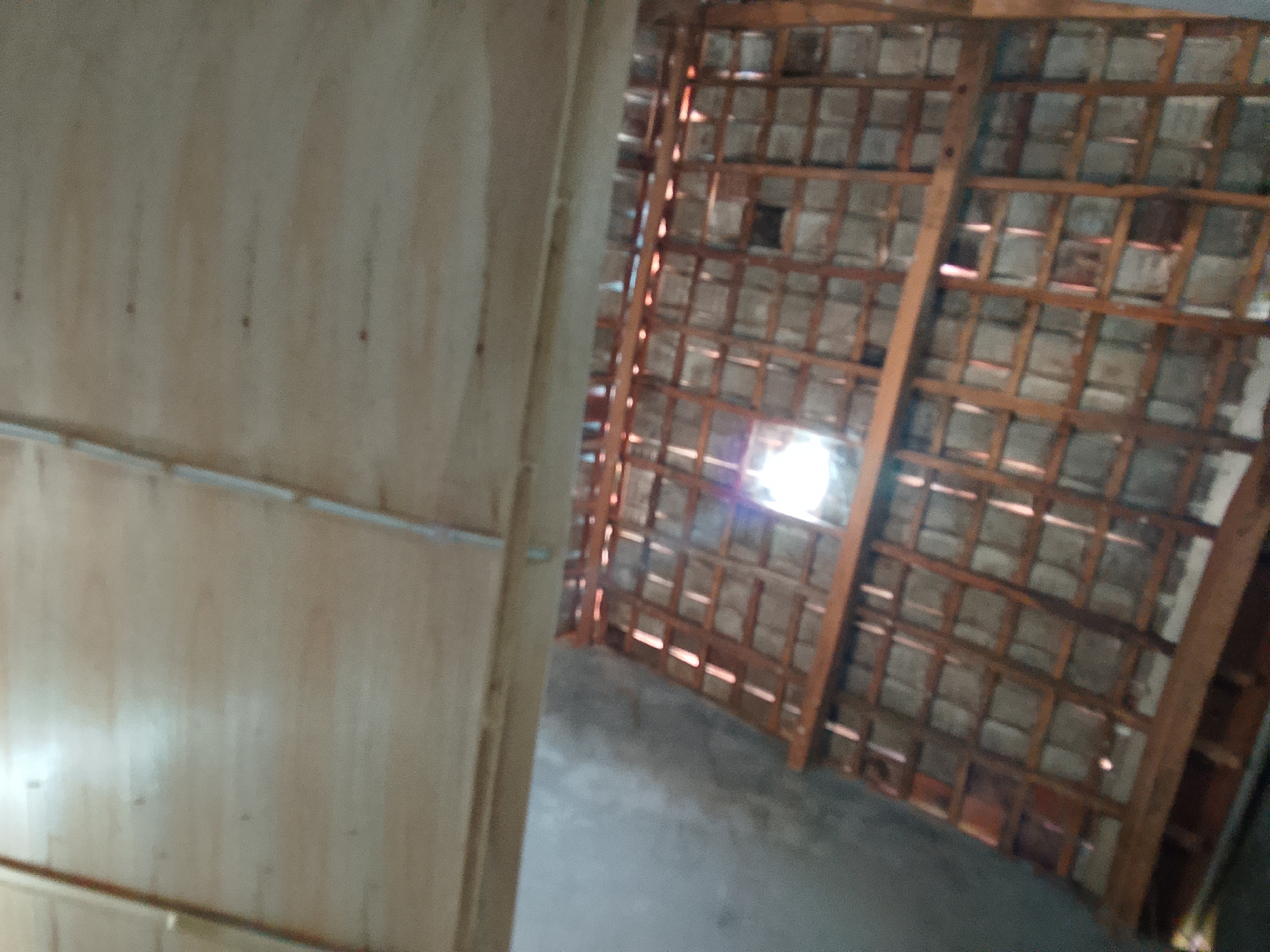 The image depicts the interior of a structure with a wooden ceiling framework supporting roof tiles. The ceiling is partially visible and appears to be made of interlocking tiles. Light filters through gaps in the ceiling, creating bright spots. To the left of the frame, a wall constructed of light-colored wooden panels is visible, with horizontal lines suggesting planks or joins. The floor is a plain gray surface, likely concrete. The overall impression is of a simple, perhaps unfinished or rural, construction. No people or specific objects of interest are discernible. This scene is located in Besuki, Indonesia.