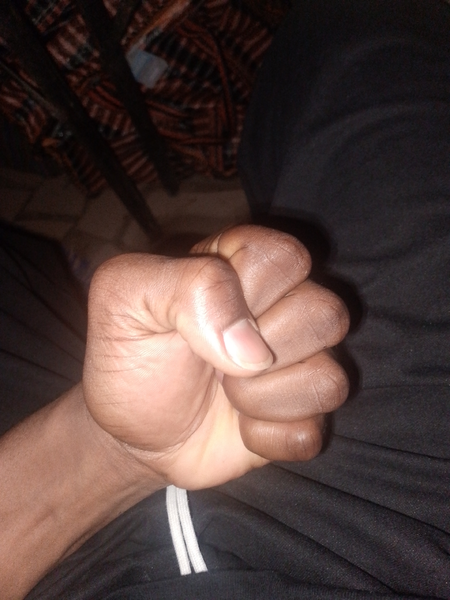 A close-up, slightly angled shot shows a person's closed fist, with dark skin, resting on dark fabric. The background features a blurry, out-of-focus pattern of red, black, and white lines, suggestive of fabric or a blanket. The lighting appears dim. The image was taken in Ipetumodu, Nigeria.