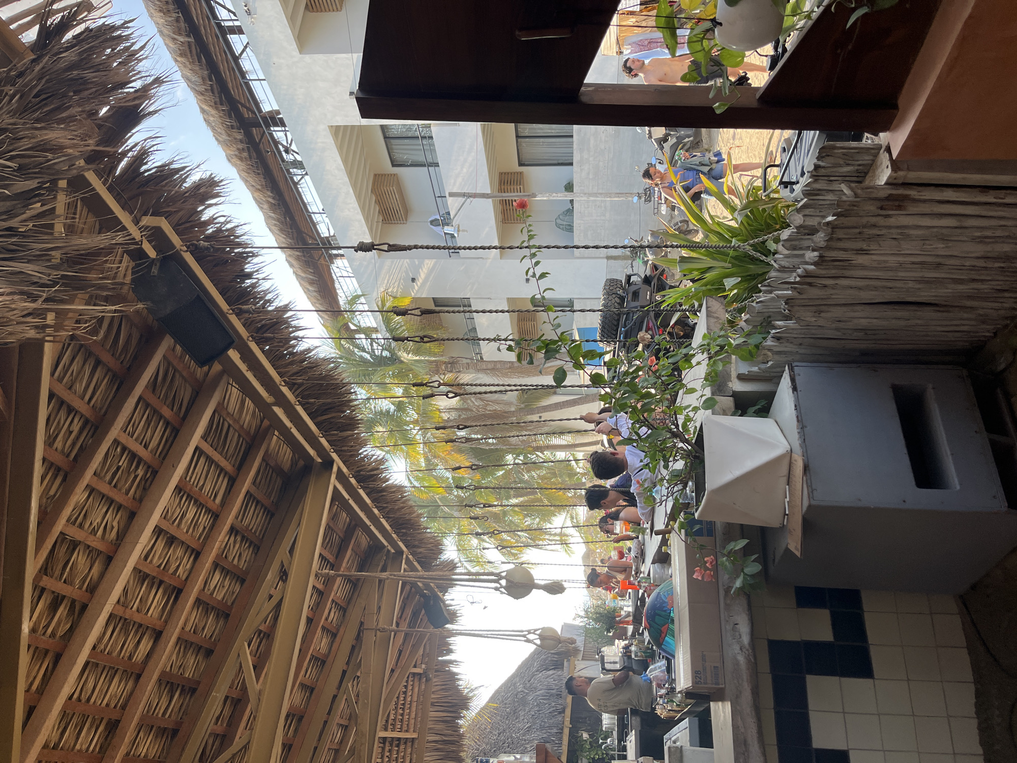 The image shows an outdoor restaurant in Brisas de Zicatela, Mexico. A thatched roof structure covers the restaurant area. A group of people are seated at tables, appearing to be dining. Outside the immediate restaurant area is a view of a building and a street where people are walking. A vehicle is also visible. The environment includes palm trees and other vegetation. The scene is a mix of restaurant activity and the surrounding urban beach environment.