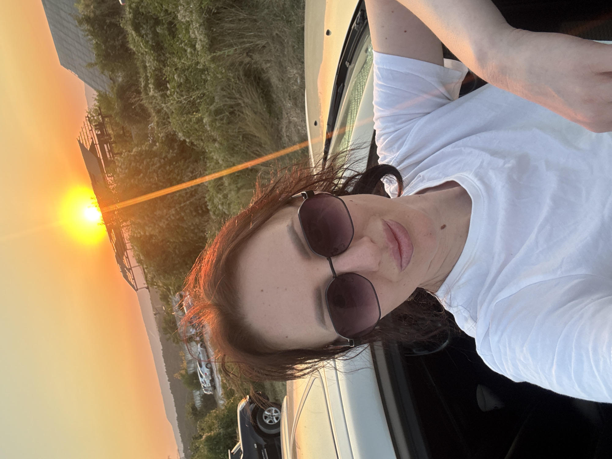 A woman with dark brown hair and sunglasses is taking a selfie in front of a sunset. She is wearing a white t-shirt and leaning against a white car. The background shows a hilly landscape with some buildings and parked cars. The sun is setting behind a structure with a slanted roof, casting a warm glow over the scene. The location appears to be outdoors, possibly in a rural or suburban area.