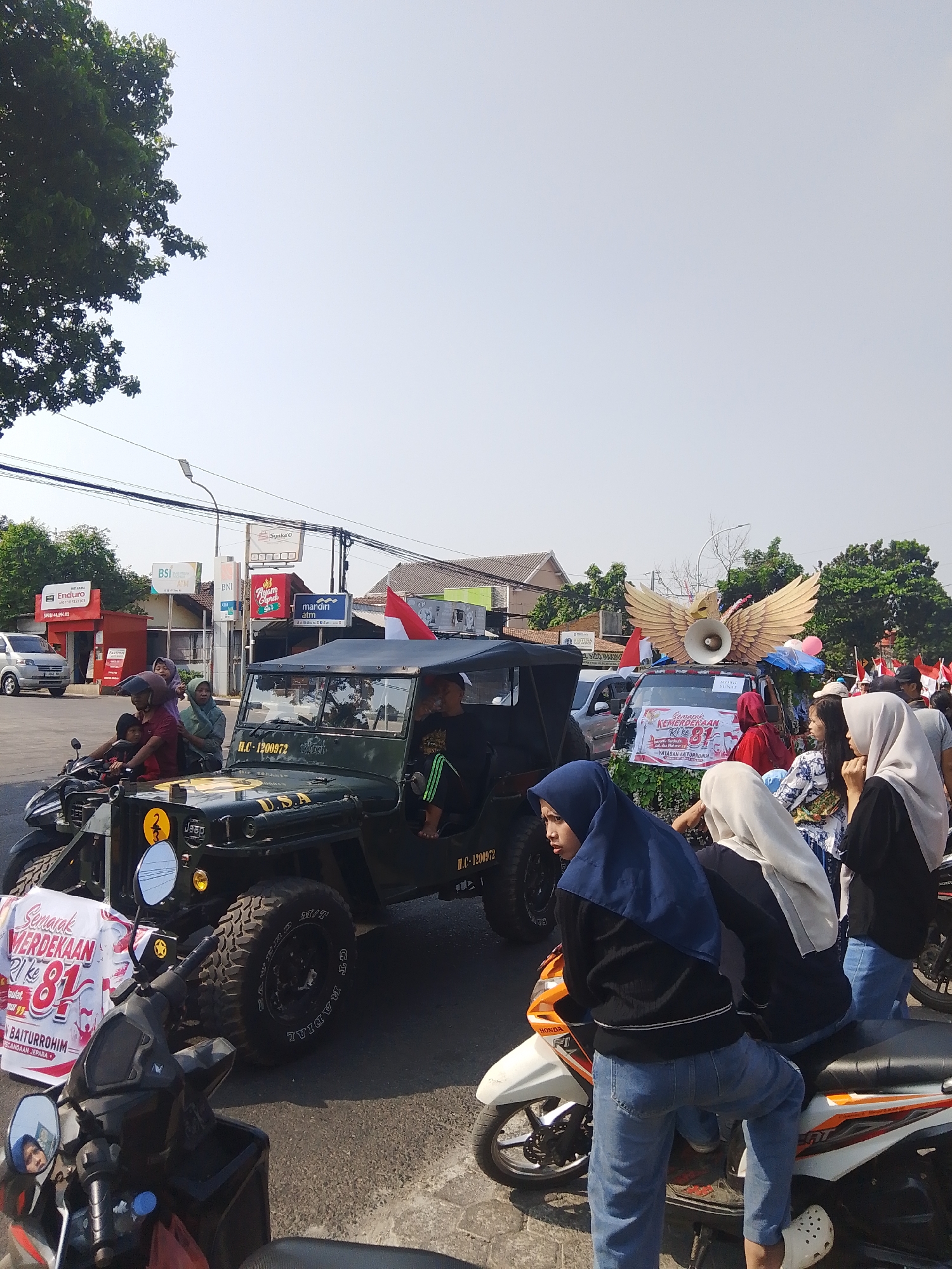 In Rejosari, Indonesia, a parade is underway on a clear, sunny day. The foreground features several motorcycles and a vintage olive-drab Jeep, adorned with a "USA" decal and bearing the license plate "H.C.-1200972." A banner celebrating "Semarak Kemerdekaan RI ke 81" (Festival of Indonesian Independence 81) is displayed prominently near the Jeep. Several individuals, including women wearing hijabs and children, are visible on and around the vehicles. In the background, more participants and spectators line the street, with Indonesian flags waving. Commercial buildings, including ATM outlets for BNI, Mandiri, and Syariah, are situated along the roadside, indicating an urban or semi-urban setting. The event appears to be a community celebration, possibly related to national holidays, with a festive atmosphere.