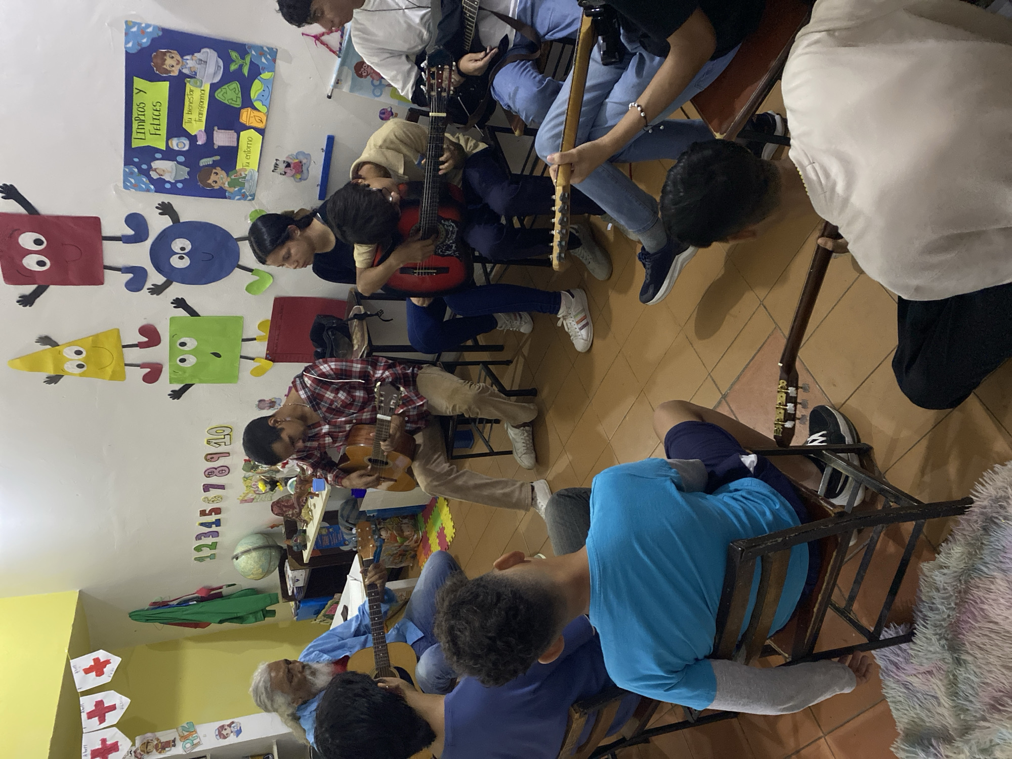 In Tinaquillo, Venezuela, a group of approximately eight people are seated in a circle, playing guitars in what appears to be a classroom. The room has educational posters and decorations on the walls. A man with a white beard is also playing a guitar. The people appear to be of varying ages.