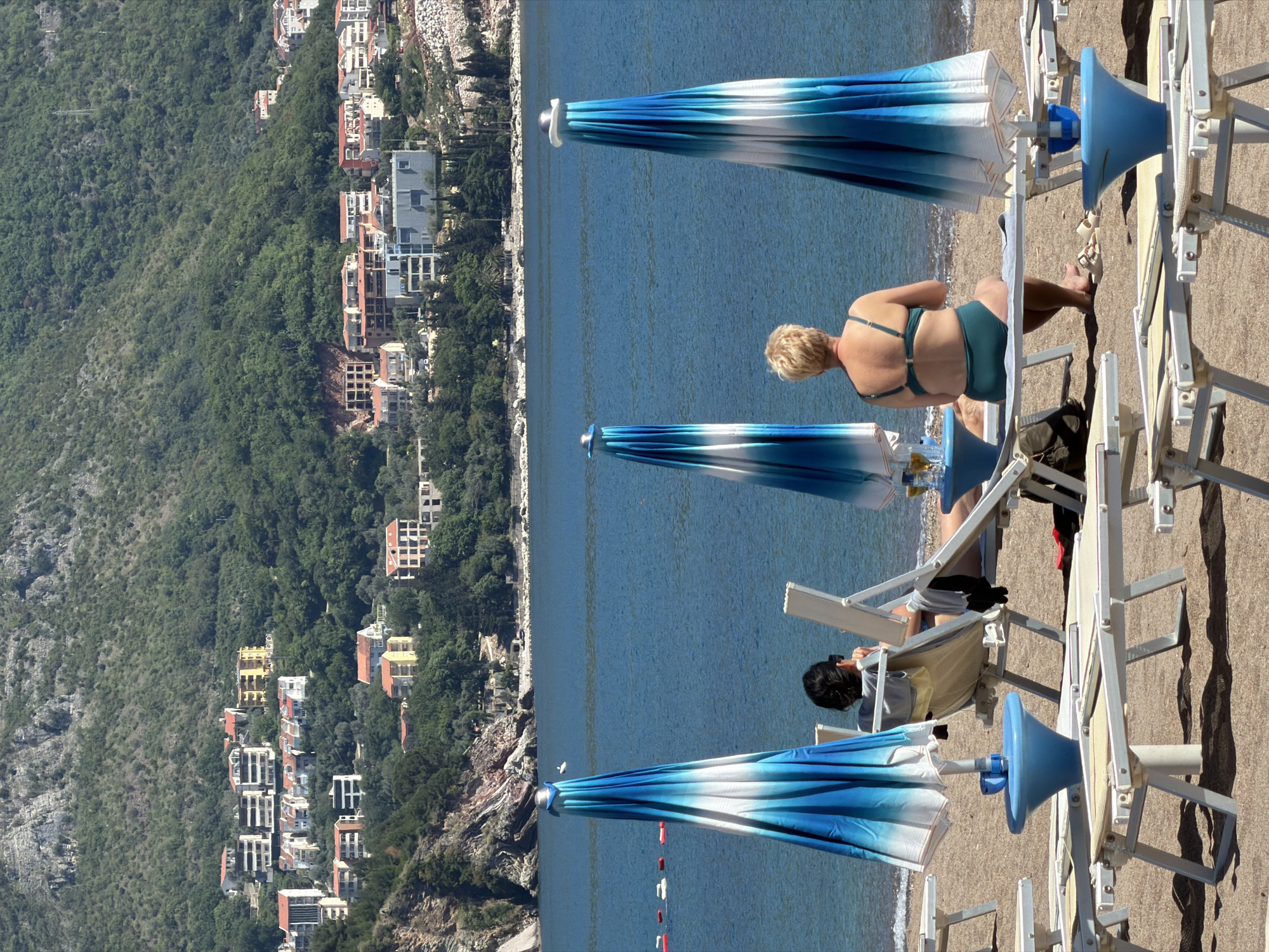 The scene presents a picturesque beach setting in Bečići, Montenegro, juxtaposing the natural landscape with human leisure.
In the foreground, three beach umbrellas, alternating blue and white, offer shade to sunbathers on white beach chairs. A woman with short blonde hair, clad in a dark green bikini, relaxes on one of the chairs. Nearby, another individual with dark hair sits on a chair, absorbed in their smartphone.
The backdrop showcases the Adriatic Sea, its calm blue surface extending to the horizon. Adjacent to the sea, the urban development of Bečići is nestled against a verdant mountain range, the structures of houses and buildings contrasting with the natural greenery. The composition highlights the blend of tourism and local life along the Montenegrin coast.
