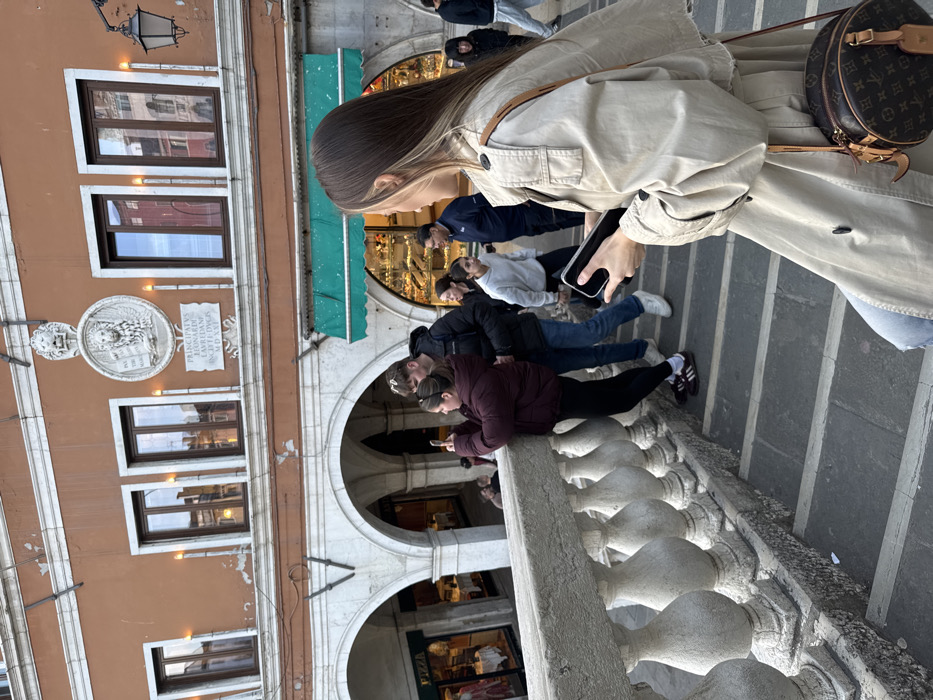 The image captures a detailed street scene on or immediately adjacent to the Rialto Bridge in Venice, Italy. The background features a reddish-orange building facade with white architectural trim, displaying three arched windows. Centrally positioned on this facade is a large circular stone relief depicting the Winged Lion of St. Mark holding a book, surmounted by a smaller lion head. Below the circular relief is a rectangular stone plaque inscribed with "PRINCIPATU LEONARDO LAUREDANO IN MDXXV" (During the Principate of Leonardo Loredan in 1525). Decorative, elongated light fixtures are mounted between the windows. Below the facade, a series of white stone arches house various commercial establishments; one visible on the left is a "PIZZA" shop, while another on the right features a green awning and a display window filled with various items.
In the foreground, to the right, a female is visible from the back, wearing a light-colored trench coat and carrying a brown, circular Louis Vuitton monogrammed shoulder bag. She has long brown hair with prominent blonde highlights and holds a smartphone in her right hand, positioned as if interacting with the device. To the left, a sturdy grey stone balustrade, featuring multiple balusters, runs parallel to the viewer, indicating an elevated walkway or bridge.
In the mid-ground, on a set of descending stone steps, a female wearing a dark maroon puffer jacket and black leggings leans against the balustrade, holding a smartphone horizontally, appearing to photograph or video an unseen subject to her left. Beside her, a male in a dark puffer jacket, blue jeans, and white sneakers stands, looking in the same general direction. Further in the background, several other individuals are present on the steps and beneath the arches, including a male in a dark blue polo shirt and a female in a light grey hoodie, engaged in walking or standing. The lighting suggests overcast daylight conditions.