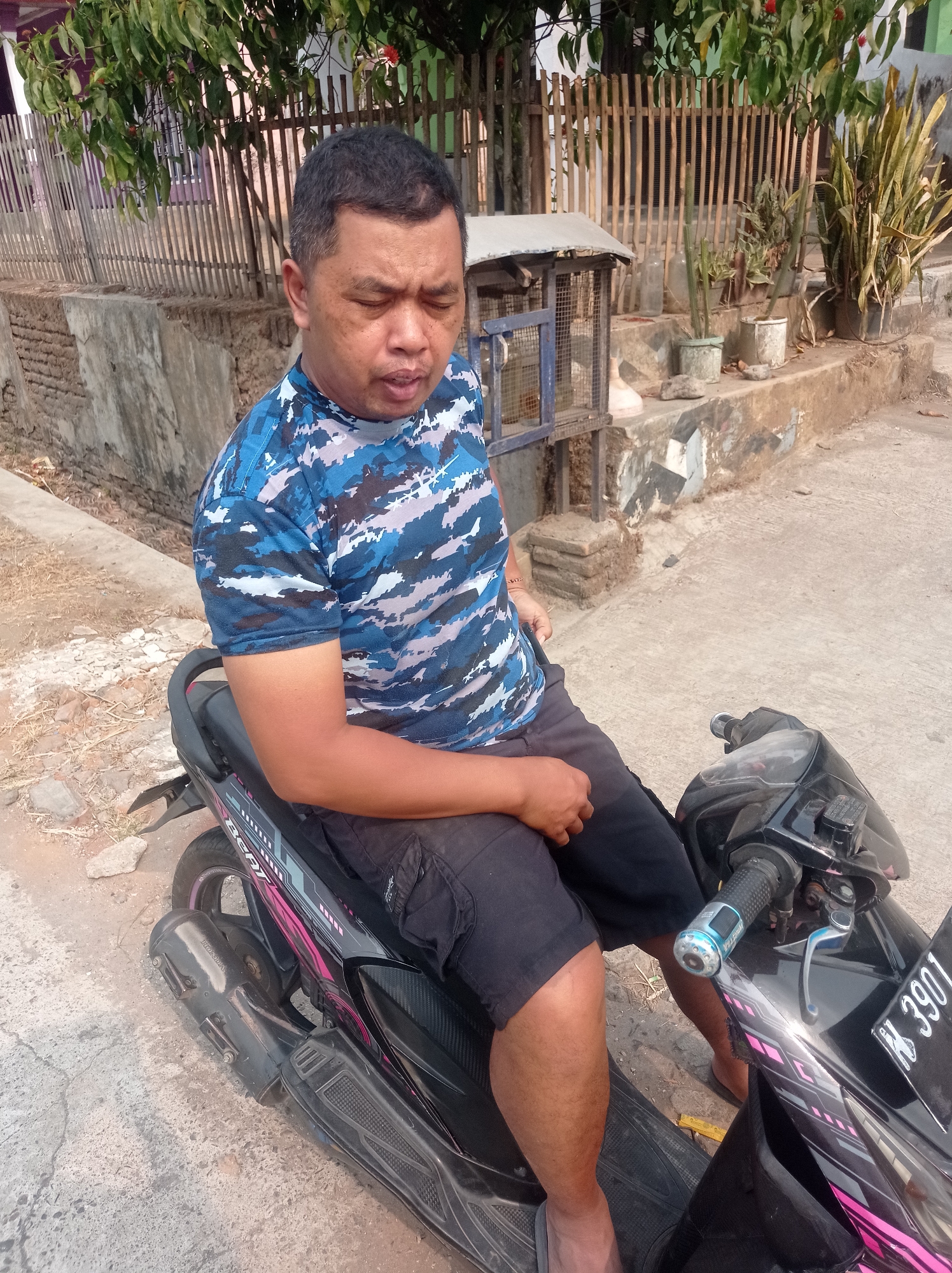 A man is sitting on a black scooter in what appears to be a residential area in Purwosekar, Indonesia. The man is wearing a blue and grey camouflage t-shirt and black shorts. He has his eyes closed and his head tilted down. The scooter has pink and grey decals on the side and a license plate that reads "N 3901". In the background, there is a wooden fence, a concrete wall, and some potted plants. There is also a small cage with a blue frame and wire mesh. The ground is concrete and dirt. The lighting suggests it is daytime.