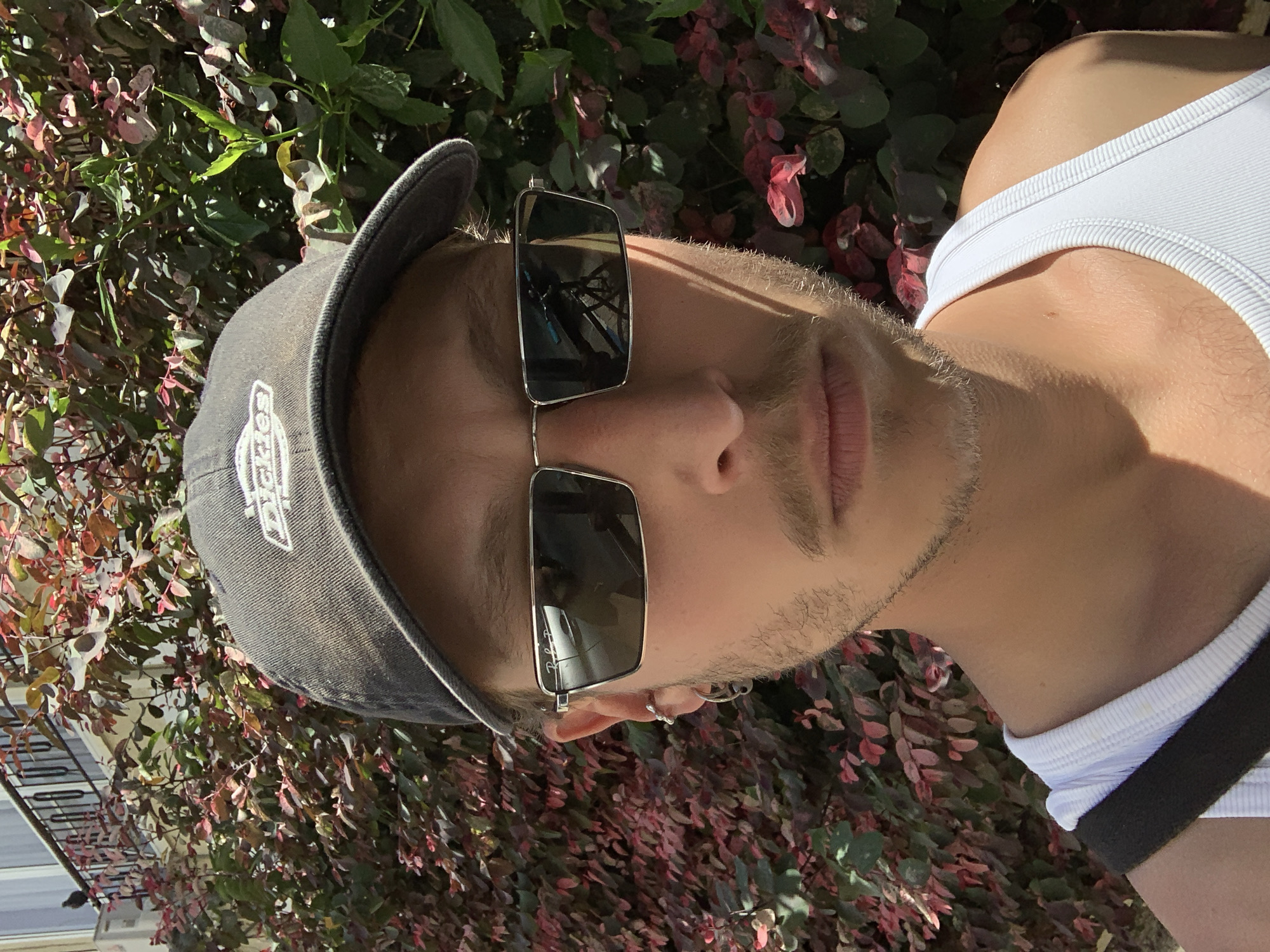 A medium shot depicts a person wearing a white tank top, a gray baseball cap, and sunglasses. The person is positioned in front of a dense bush with reddish-purple foliage. The lighting suggests it is daytime. The location appears to be outdoors, possibly in Tel Aviv, Israel, although there are no immediately distinguishing landmarks confirming it.