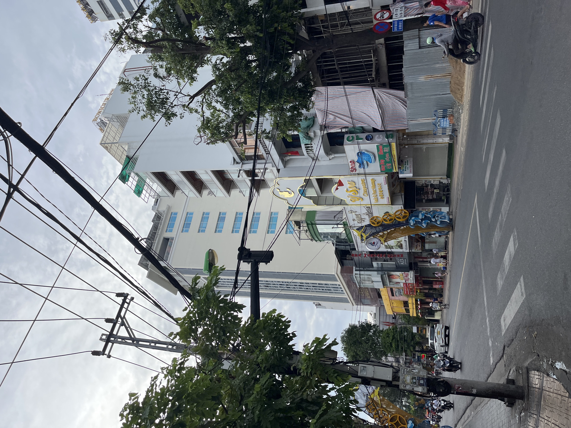 This is a street scene in Nha Trang, Vietnam. The image captures a road with pedestrian crossings and traffic, primarily consisting of motorcycles. Several multi-story buildings line the street, featuring a mix of modern architecture and older shopfronts. Overhead, a dense network of electrical wires and poles dominates the sky, a common sight in Vietnamese cities. Some buildings display signage for businesses like "Bonjour Hotel," "Y Spa," and "Crocs Shoes." People are visible walking on sidewalks and crossing the street, while others ride motorcycles. The environment appears to be urban, with construction materials, such as corrugated metal sheeting, visible in front of one building. The sky is overcast, suggesting a cloudy day.