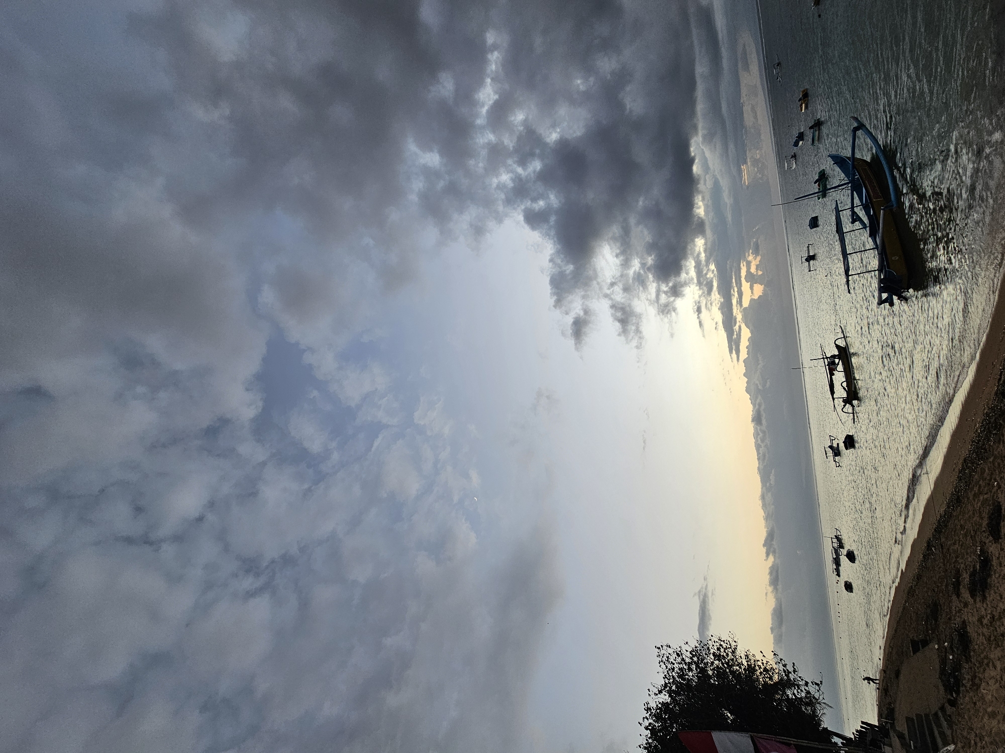 The image depicts a serene beach scene at dawn in Denpasar, Indonesia. The sky is a canvas of soft blues and grays, with streaks of pale yellow near the horizon, indicating the rising sun. Dark, heavy clouds loom in the upper right portion of the frame, contrasting with the lighter clouds scattered across the rest of the sky. A crescent moon is visible in the upper left quadrant of the sky. The calm sea stretches out to the horizon, dotted with several traditional Balinese fishing boats, known as jukungs. One prominent jukung, yellow and blue, is closer to the foreground and appears to be anchored. The sandy beach occupies the lower left of the image, with a small section of stairs leading up from the shore. A lone figure stands on the beach near the water's edge. To the far left, the dark silhouette of a tree and a flag with red and white bands are partially visible. The overall atmosphere is peaceful and tranquil, capturing the quiet beauty of a coastal sunrise.