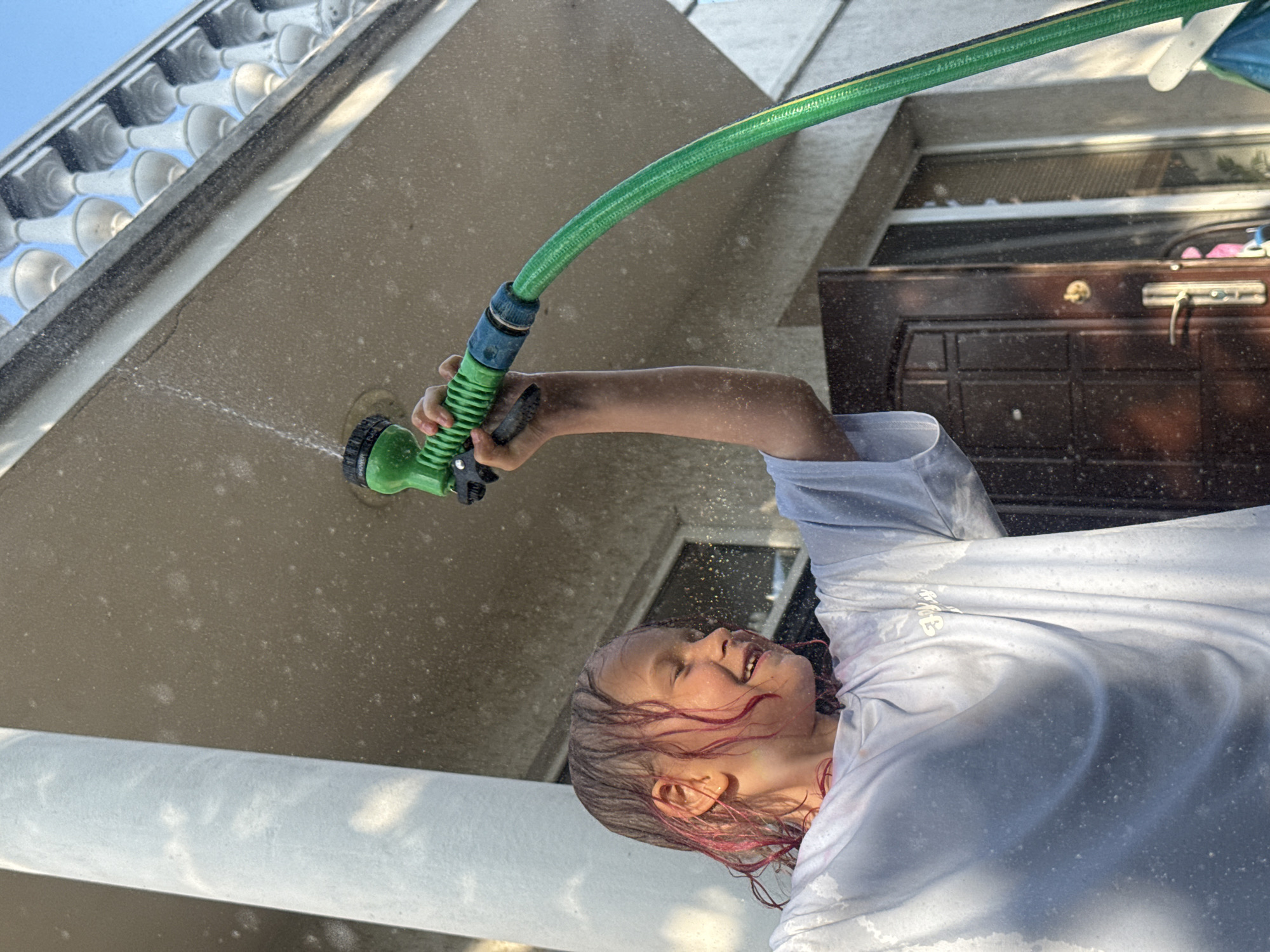 An outdoor, eye-level image depicts a young child, likely female, with wet, light-colored hair featuring reddish or pink streaks. The child's eyes are closed, and a broad smile is visible on her face. She wears a light-colored, long-sleeved t-shirt that is thoroughly saturated with water, clinging to her form. Her left arm is raised, holding a green garden hose nozzle which is actively spraying water upwards and towards her. The nozzle has visible text, including "SHOWER" and "FLAT". Numerous water droplets are suspended in the air around the child, the hose, and the background.
The setting is an exterior residential area. To the left, a large, light-colored architectural column stands. Above and to the right, a white balustrade with several decorative balusters is visible against a clear, light blue sky. The background consists of a light-colored building wall, which also appears wet or has water droplets adhering to it. In the lower right background, a dark brown, paneled door with a brass-toned handle and lock mechanism is partially visible, adjacent to a window fitted with blinds.