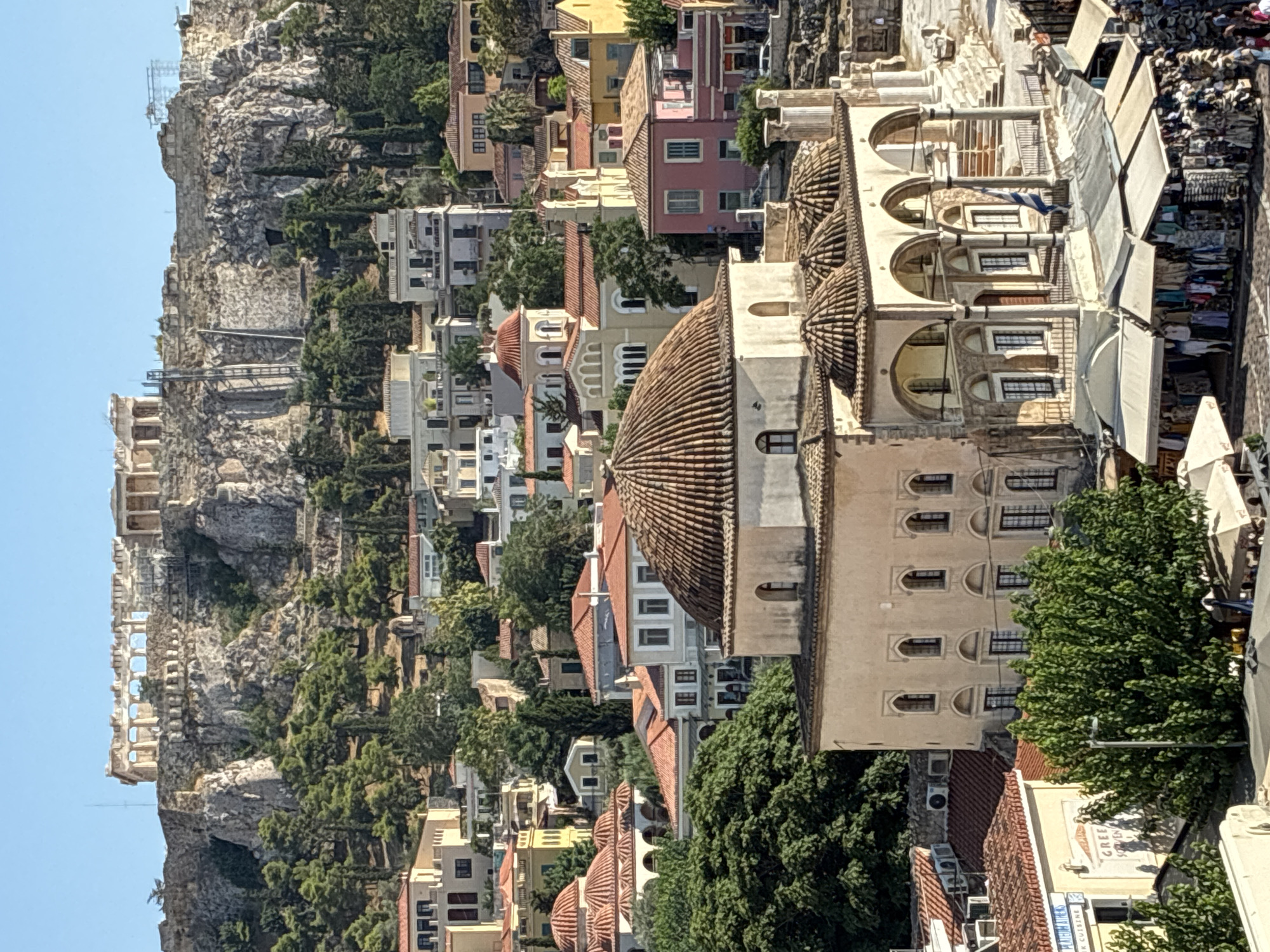 The image captures a view of the historic city of Athens, Greece, with the Acropolis perched atop a rocky hill in the background. In the foreground, a domed building with arched porticoes, likely a mosque or historical bathhouse, stands prominently. Below this structure, several market stalls are visible, displaying clothing and souvenirs. The surrounding area is filled with traditional Athenian architecture, characterized by white and pastel-colored buildings with terracotta roofs, interspersed with lush green trees. The clear blue sky suggests a sunny day.