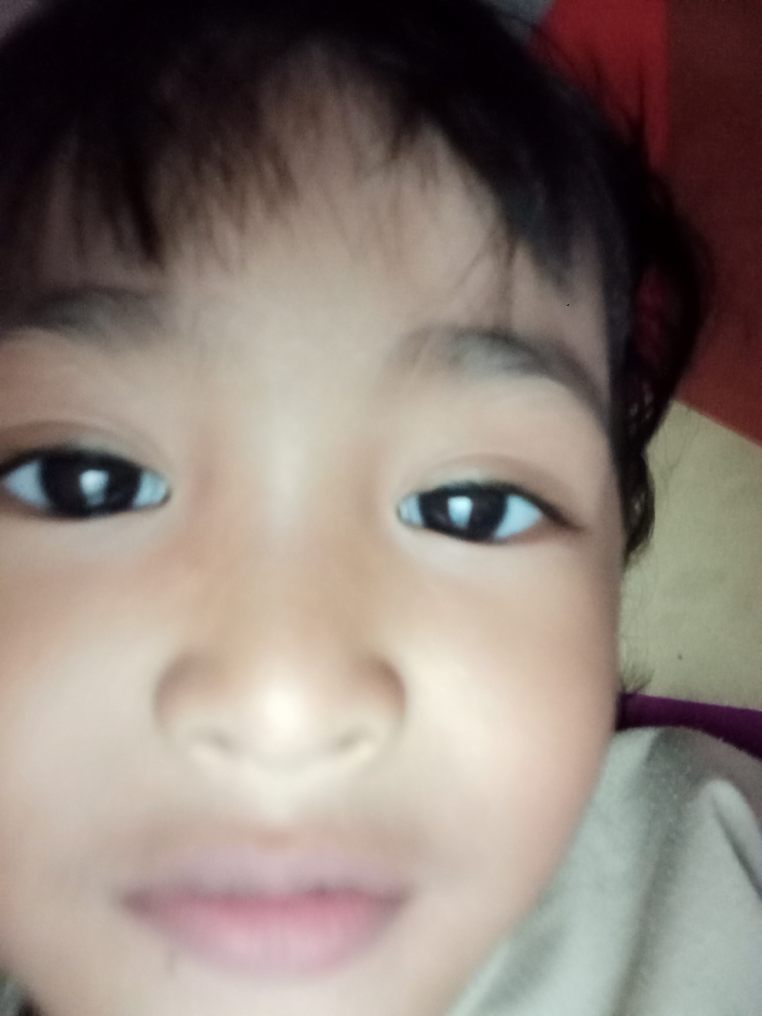 The image is a close-up, slightly blurry photograph of a young child's face, viewed from the front. The child has dark hair, dark eyes, and fair skin. The lighting is soft, and the background is out of focus, appearing as indistinct shapes of red and yellow. The child is wearing a light-colored garment. There are no identifiable landmarks or context to suggest a specific location within Bloro, Indonesia.