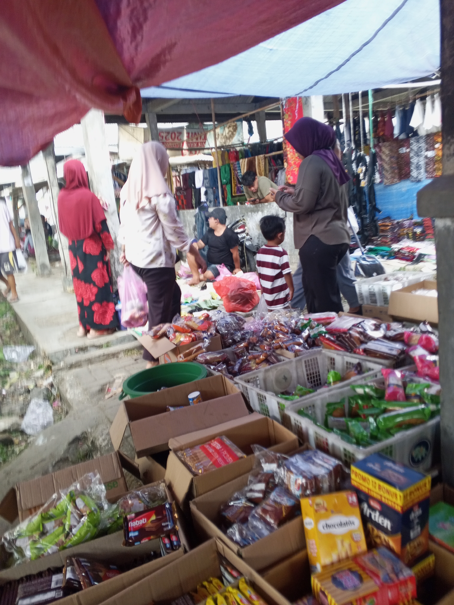 The image depicts an outdoor market stall in Serang, Indonesia, likely a traditional market given the setting and goods. The scene is crowded with goods displayed in cardboard boxes and plastic crates. Various food items, primarily packaged snacks and biscuits, are visible, including brands like "Nabati" and "Arden." Several individuals are present, including women wearing hijabs, suggesting adherence to Islamic cultural norms prevalent in Indonesia. One woman in a red and black floral dress stands near the foreground, while another in a beige hijab carries a pink plastic bag. A child in a striped t-shirt stands to the right, facing away from the camera. A man is seated in the background, possibly a vendor. The market is covered by tarps, providing shade from the sun. The overall impression is of a bustling marketplace scene, typical of local commerce in Southeast Asia.
