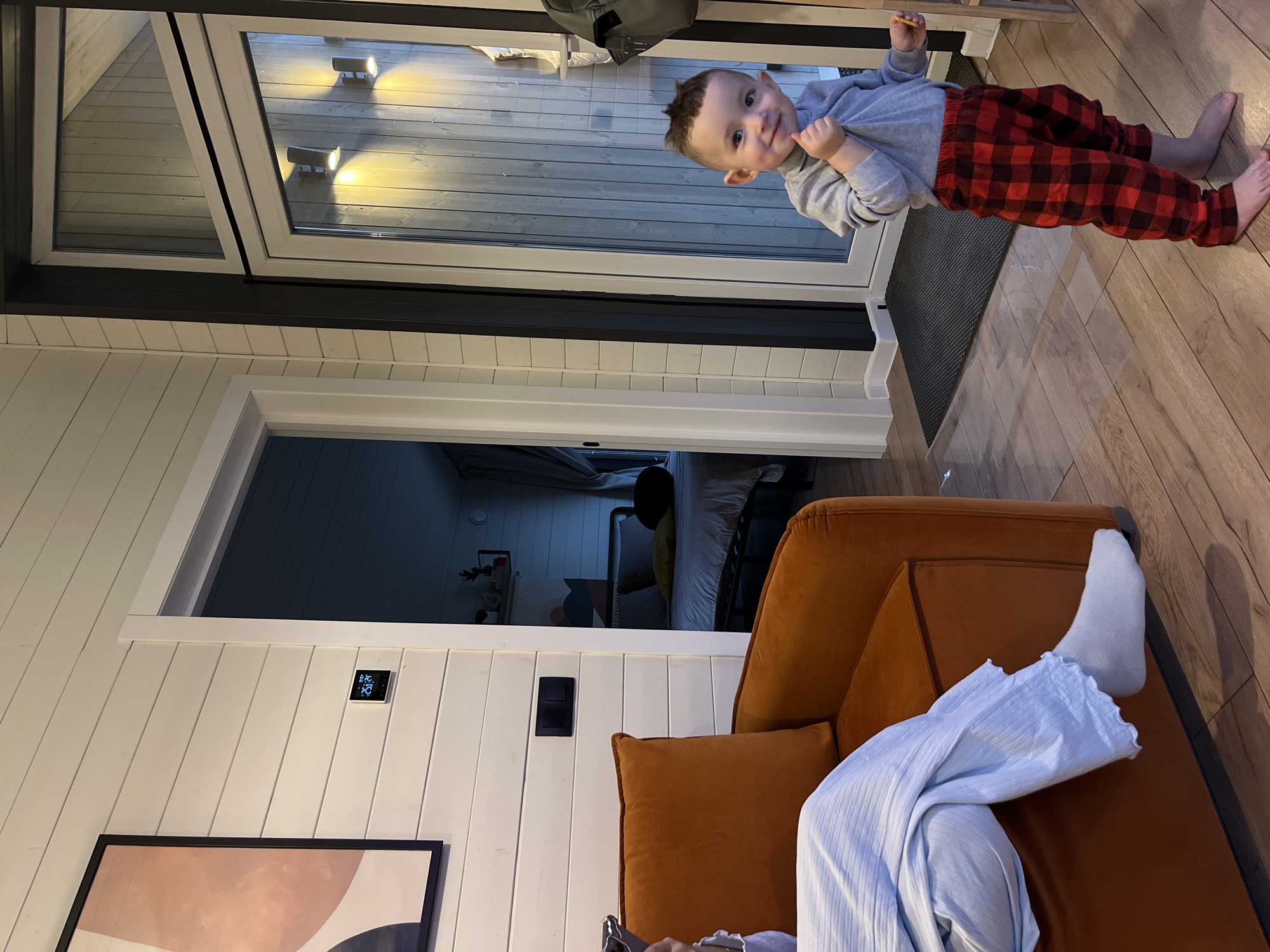 An indoor scene captured in Litvinovo, Russia, features a young child and a partially visible adult, set against a modern interior with outdoor elements. The image is taken from a slightly elevated perspective, looking down.
In the foreground, a smiling young boy with short, dark hair and blue eyes stands on a light-colored wooden floor. He wears a grey long-sleeved shirt and red and black plaid pajama pants, with bare feet. His left hand is raised towards his chest, and his expression is joyful and playful, looking directly at the camera. He is positioned near a large glass door or window, which offers a view to an outdoor deck.
Beyond the glass, the deck is illuminated by two wall-mounted lights that cast a warm, yellowish glow on the wooden planks, suggesting it is either dusk or nighttime outside. A dark door mat is visible on the floor near the threshold of the glass door.
To the right, an orange sofa occupies a significant portion of the frame. The legs of an adult, clad in light blue ribbed pants and white socks, are visible resting on the sofa, indicating someone is relaxing.
The interior walls are covered in white horizontal paneling. A framed piece of art, displaying abstract shapes in muted tones (pink, black, white), hangs on the lower left wall. Above it, a black digital display unit, likely a thermostat or smart home control, is mounted. The screen shows numbers like "22" and "20" along with degree symbols and other small icons.
Through an open doorway in the background, another room, possibly a bedroom, is visible. It appears dimly lit, with a bed and dark headboard noticeable. The overall impression is one of a cozy, modern living space.