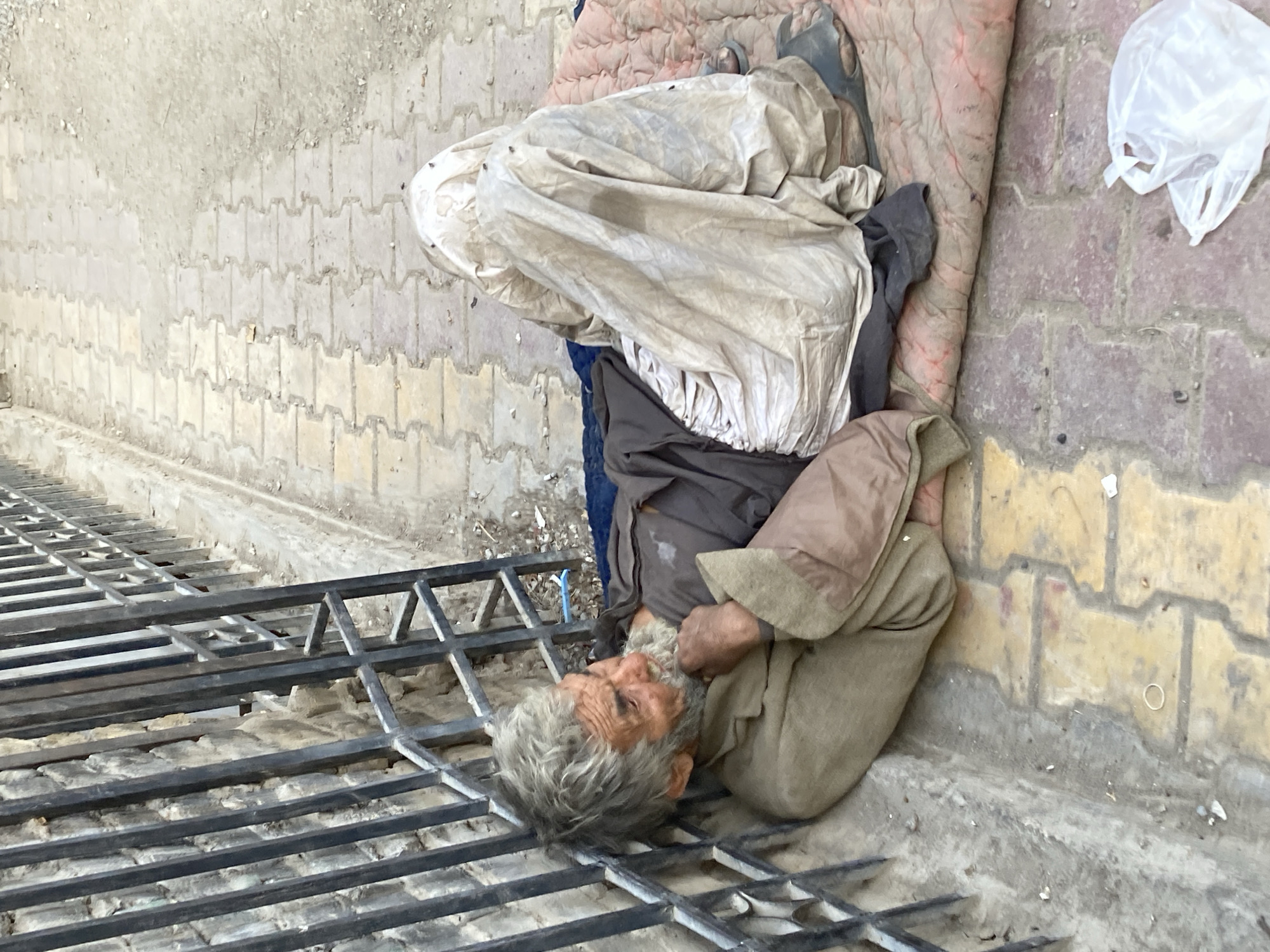 A disheveled elderly man with gray hair and beard lies on a metal grate beside a sidewalk. He is partly covered with a white cloth, blankets and a dirty brown garment. A plastic bag sits on the sidewalk. The location is Quetta City Tehsil, Pakistan.