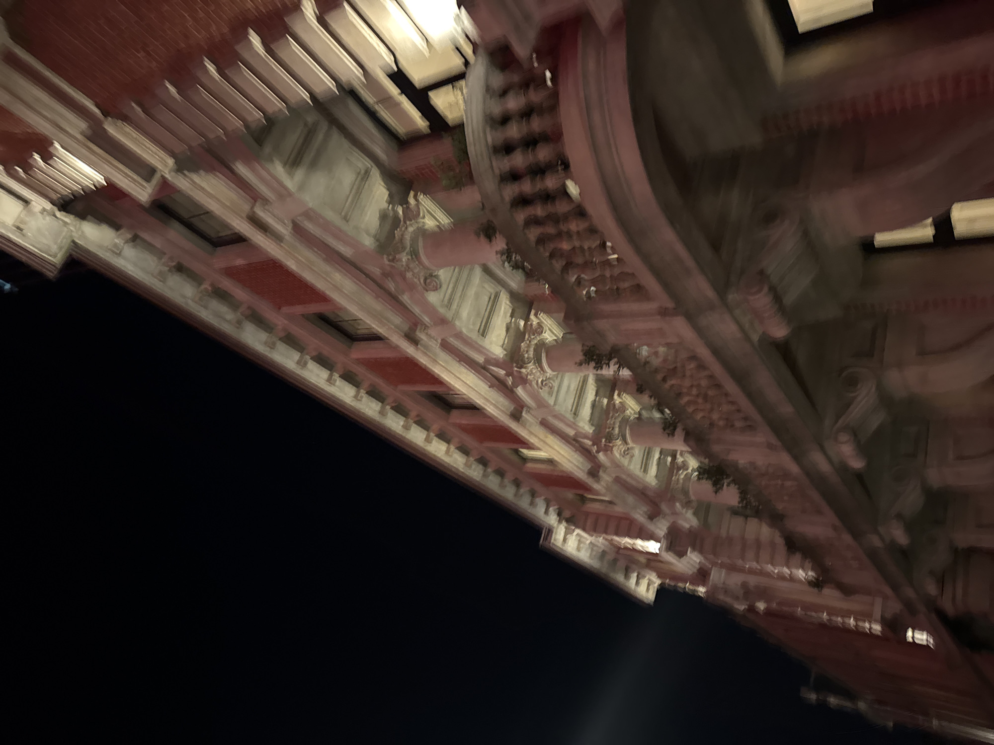 The image displays a low-angle, diagonally rotated perspective of a grand architectural structure at night, likely located in Perth, Australia. The building is illuminated by artificial lights, casting a warm, reddish-orange glow on some sections, possibly brickwork, and highlighting intricate details in white or cream stone.
The structure features multiple levels of ornate classical architecture, including prominent cornices, decorative balustrades or railings, and columns with detailed capitals. The overall design suggests a historic and significant building, possibly a public edifice like a government building, museum, or theatre, showcasing rich craftsmanship.
The scene is set against a dark, black night sky that occupies the lower-left portion of the frame, contrasting sharply with the brightly lit building. The upward perspective emphasizes the height and grandeur of the architecture. There is a noticeable motion blur throughout the image, particularly evident on the building's features, which gives it a dynamic and slightly ethereal quality. No human activity is visible, and there is no discernible text. The image focuses solely on the dramatic, illuminated architecture at night.