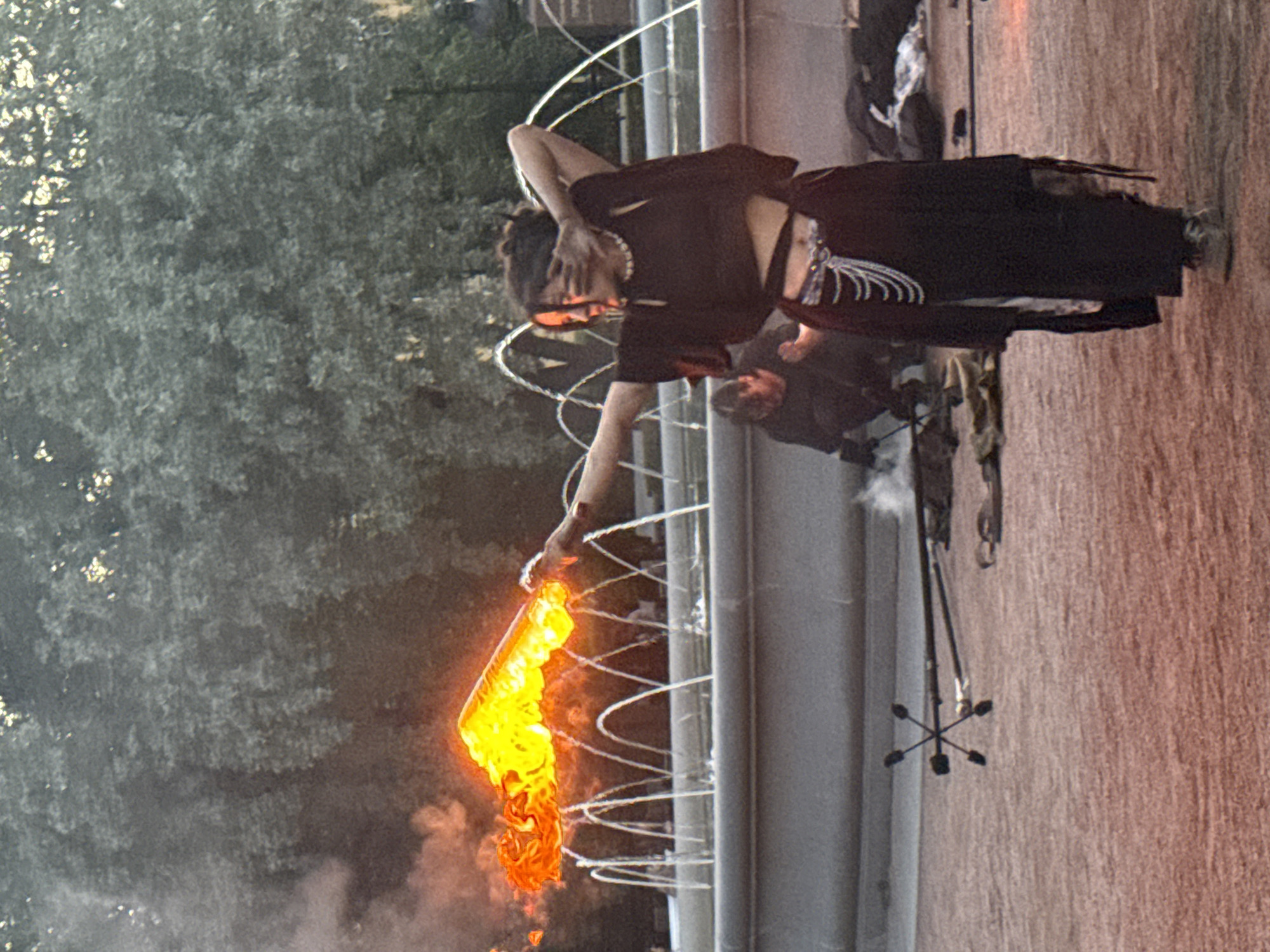 The image depicts a street performance in Saint Petersburg, Russia, likely near a fountain. In the foreground, a performer with dark hair pulled back is holding a flaming prop, possibly a staff or sword, which is emitting bright orange and yellow flames. The performer wears a black, cropped top with wide sleeves, a long, dark skirt, and a decorative beaded belt. Their right arm is extended, holding the flaming object, while their left arm is bent at the elbow, with their hand partially covering their face.
In the background, a multi-tiered fountain with arched water jets is visible. Behind the fountain, lush green trees form a dense backdrop. To the left of the fountain, a second individual is partially visible, crouched down and wearing dark clothing. The ground in the foreground is a reddish-brown gravel or dirt. A prop with four pointed ends, resembling a star, is lying on the ground in the lower left of the frame, along with other performance-related items. The overall lighting suggests it is either early morning or late afternoon, with a soft, diffused light illuminating the scene.