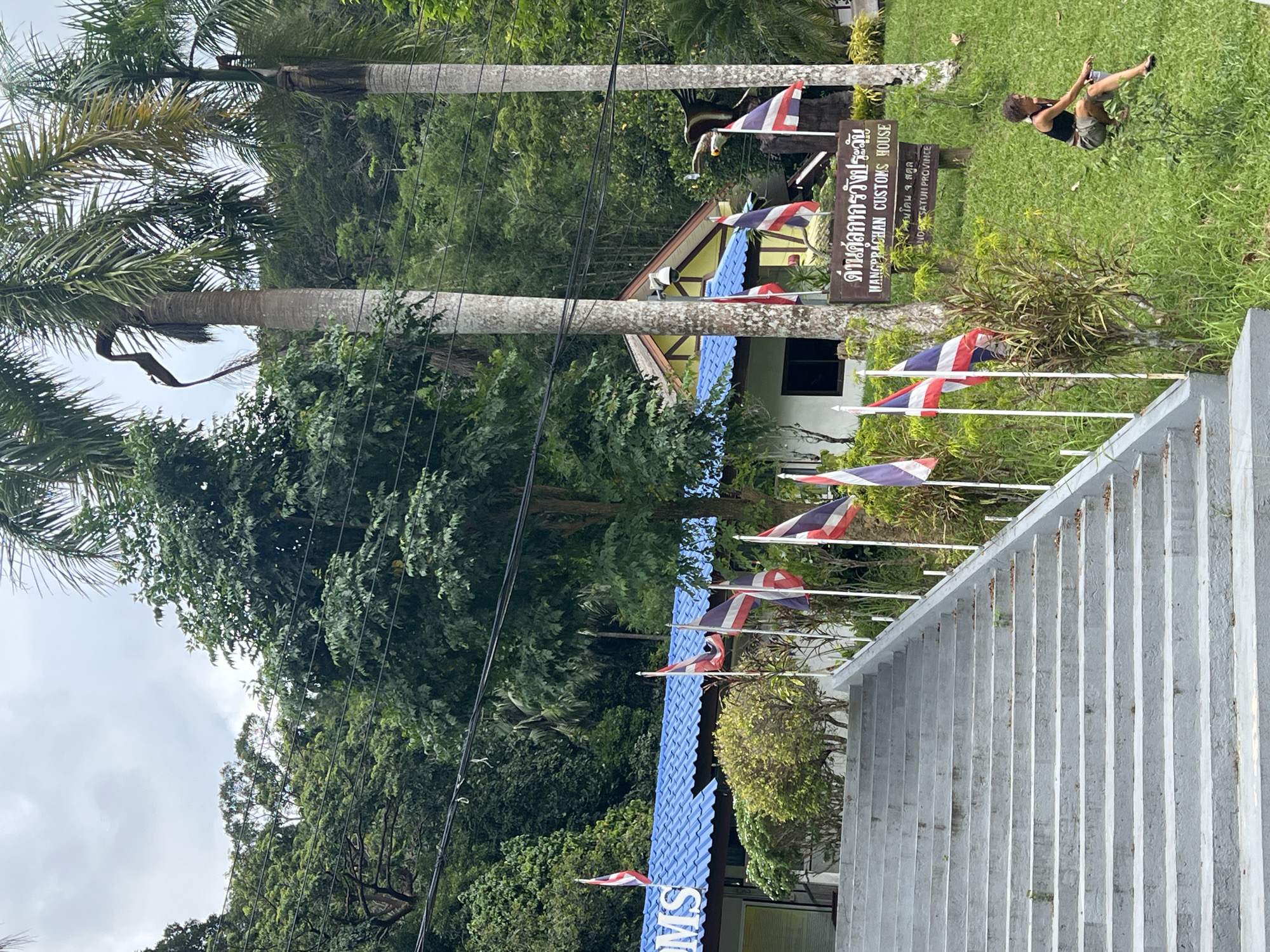 In the city of Tambon Wang Prachan, Thailand, a wide, grey concrete staircase leads up to a building with a blue tiled roof. Multiple flags, predominantly Thai flags, are planted in the ground on either side of the stairs and in front of the building. The building itself appears to be a customs house, indicated by a sign that reads "WANGPRACHAN CUSTOMS HOUSE". A person is seated on the grassy slope to the right of the stairs, near the sign, looking towards the building. The environment is lush and green, with palm trees and dense foliage surrounding the scene, suggesting a tropical setting. The sky is overcast.