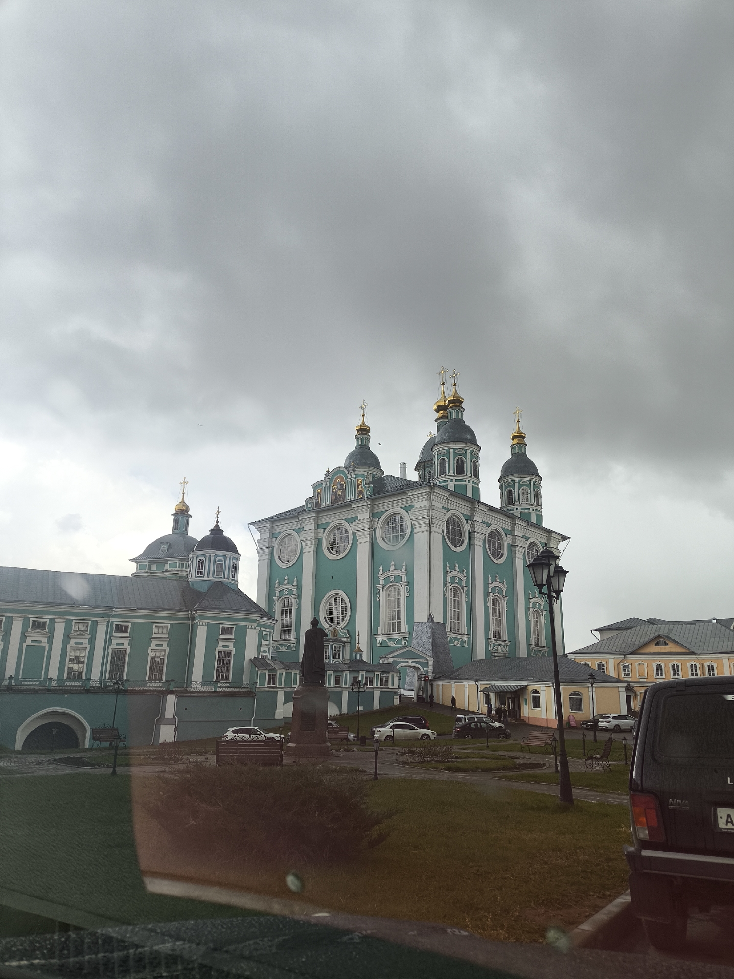 The image shows a large, elaborately decorated cathedral with multiple domes topped with golden crosses. The building is painted in shades of green and white with decorative windows. In front of the cathedral is a statue on a plinth. Several cars are parked in the square, and a black vehicle is partially visible in the foreground on the right. A street lamp stands near the edge of the green space. The sky is overcast with heavy clouds. The location is a city in an unknown region, as the location is provided with "Unknown, Unknown."