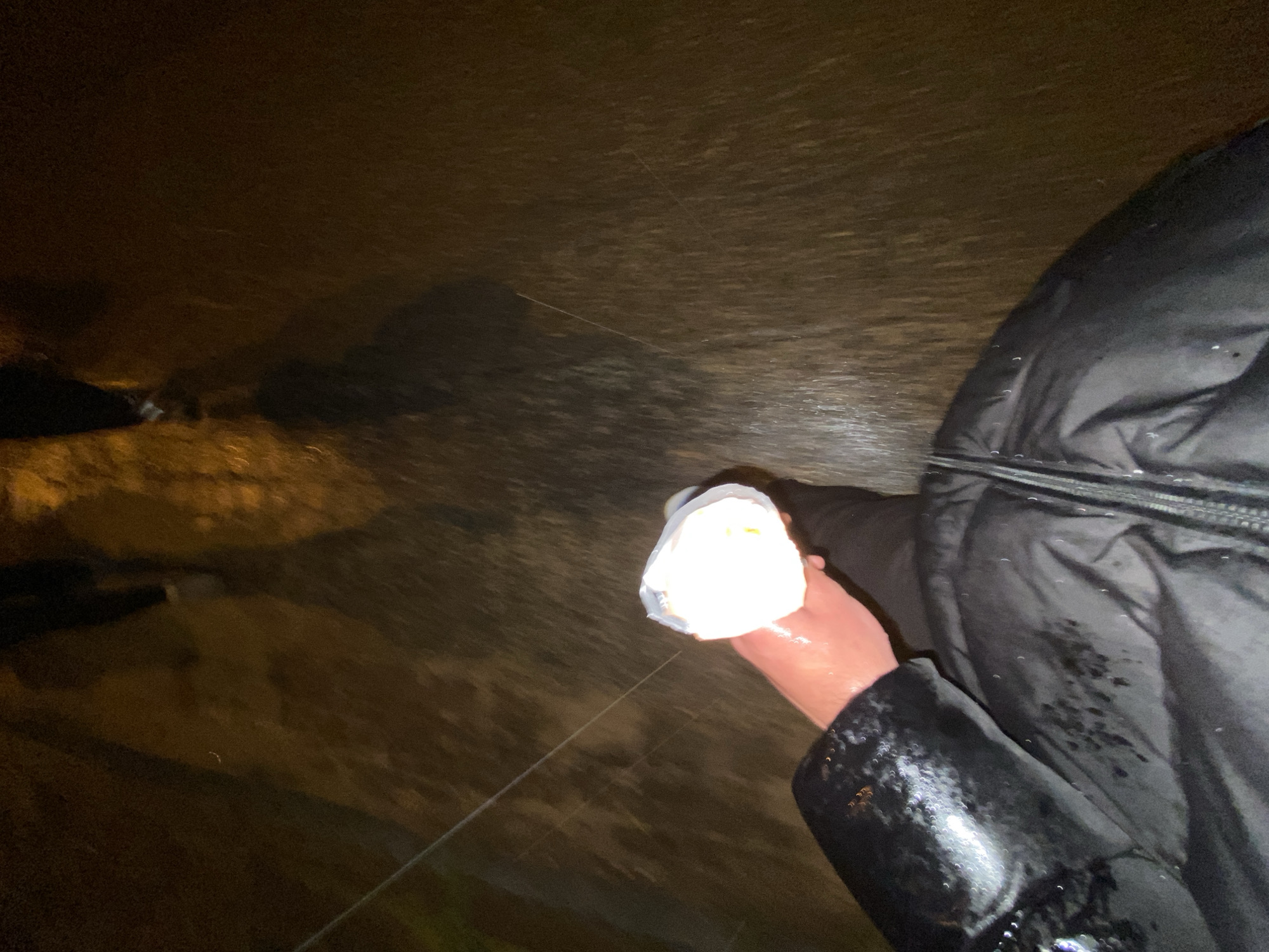 The image shows a person holding a bright, white object in their right hand. The person is wearing a black, puffy jacket that appears wet. The background is dark and blurry, suggesting a nighttime setting, potentially on a wet surface. There is a thin, light-colored line extending diagonally from the object to the left side of the frame. The photo's location is unknown. The city of Konstantinovo, Bulgaria is also unknown. The objects or environment in the images are also unknown.