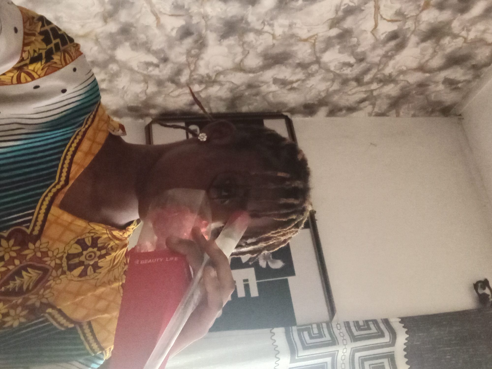 An individual is holding a red "Beauty Life" product tube. They are wearing a patterned yellow and blue garment. The background includes a framed black and white graphic, patterned wall, and ceiling. The location is potentially Shagamu, Nigeria.