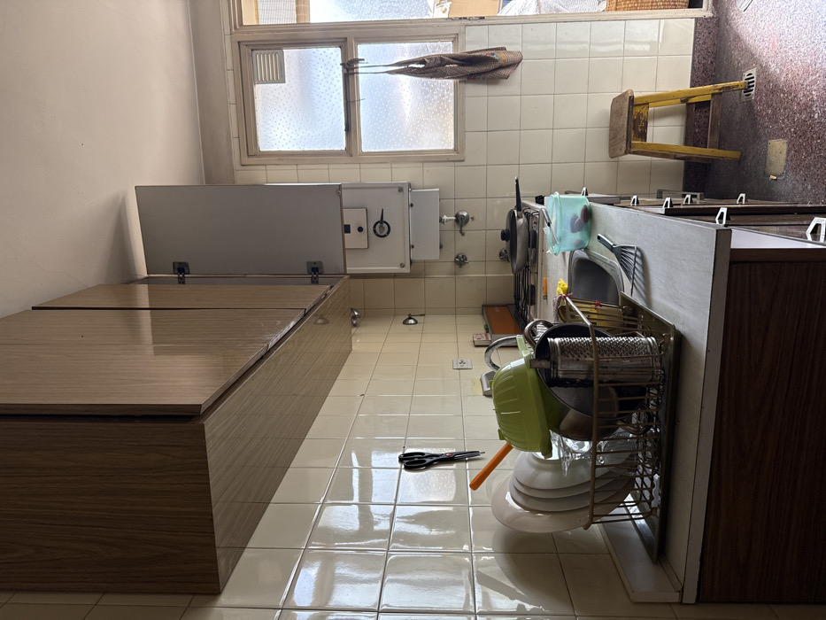 This image depicts a residential kitchen interior, characteristic of an apartment in Buenos Aires, Argentina. The scene is illuminated by natural light entering from a window on the right.
The central subject is a countertop running along the left and back walls, featuring a stainless steel sink with a faucet, and a white gas stovetop with a dark pan on one burner. To the left of the sink, a metal dish drying rack holds multiple white plates, bowls, glassware, a green plastic colander, a metal grater, and other utensils. A light blue plastic container with red contents rests near the stovetop. The wall above the countertop is covered in cream-colored square tiles, from which a pair of black scissors hangs. Above the tiled backsplash, a row of wood-grain upper cabinets extends across the top, with one door on the far right section appearing slightly ajar.
To the right of the stovetop, a wall-mounted grey cabinet with an open door reveals a white gas water heater (calefón) with visible controls. Adjacent to this, a window with white frames and translucent or frosted glass panes allows daylight to penetrate, obscuring the view outside. A small vent is present in the upper right pane of the window, from which a patterned beige apron hangs.
The floor is covered in reddish-brown mosaic or terrazzo tiles. A distressed yellow wooden stool with three visible legs stands on the right side of the frame, near the window. No people are visible, but the arrangement of objects suggests recent domestic activity.