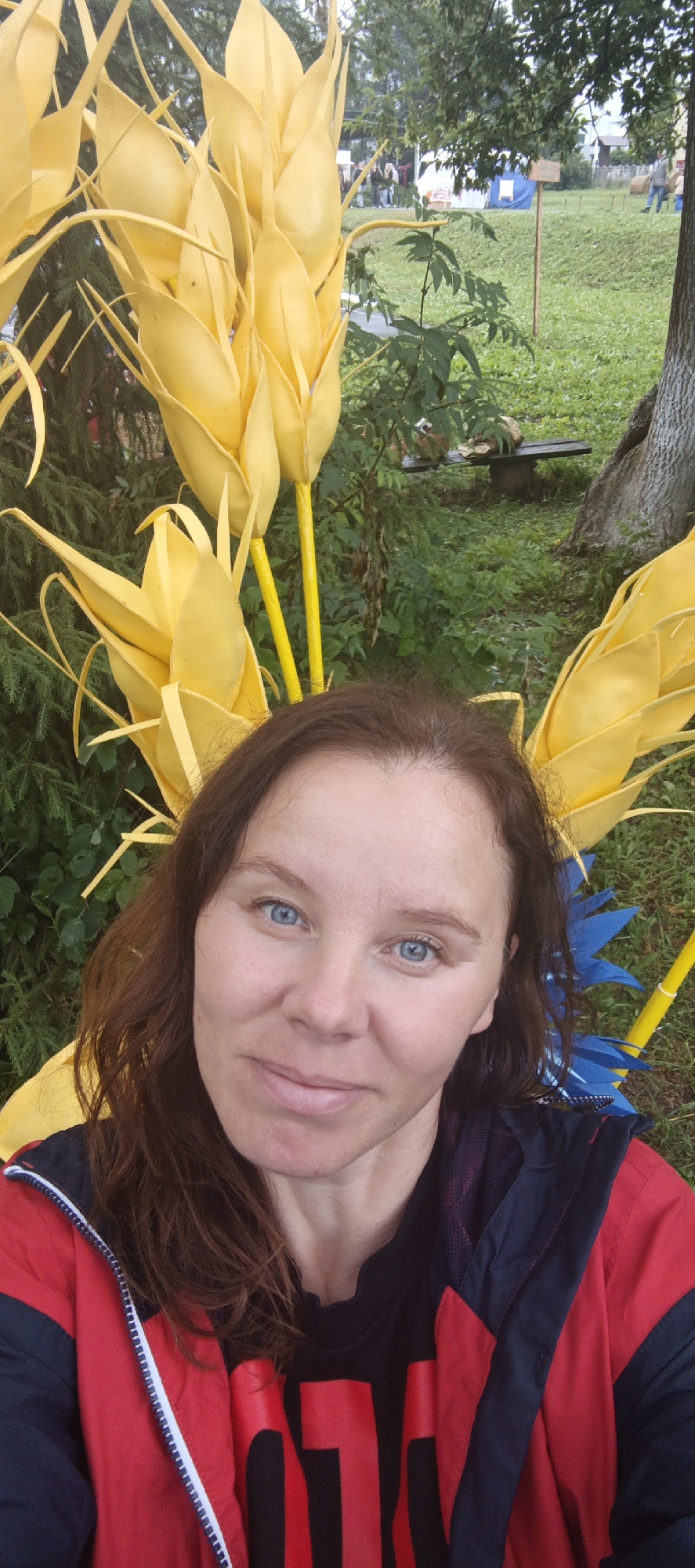A woman with brown hair and blue eyes is taking a selfie outdoors. She is wearing a red and blue jacket over a black shirt with large red lettering. She is positioned in front of large, yellow, stylized wheat stalks and some blue floral decorations. The background appears to be a grassy area with trees and some tents, suggesting an outdoor event or festival. The location is identified as Старые Быги, Russia.